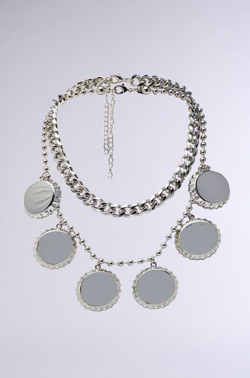 The POP THE TOP SILVER NECKLACE SET showcases a thick chain link design with faux silver, featuring scalloped edge pendants resembling bottle caps. These round, flat charms are evenly spaced and reflectively ornate against a simple backdrop.