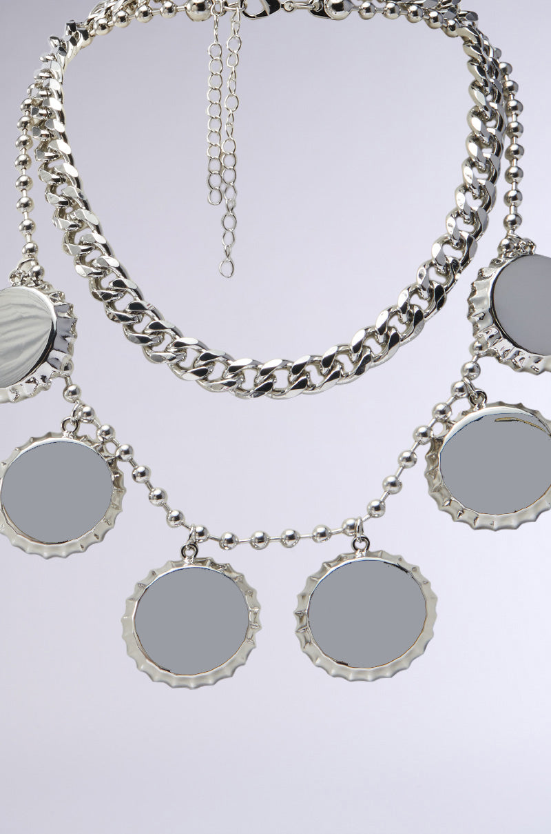 The POP THE TOP SILVER NECKLACE SET features a faux silver chain link design with large round reflective discs akin to bottle cap charms. Scalloped edges add flair, and small beads enhance the decor against a plain gray backdrop.