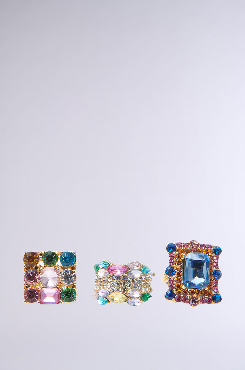 The AKIRA Label's THREE WISHES RING SET is shown on a light grey backdrop, featuring three ornate rings: one with pink, green, and amber stones; another with a rainbow of gems; and the third highlighted by a large blue stone.