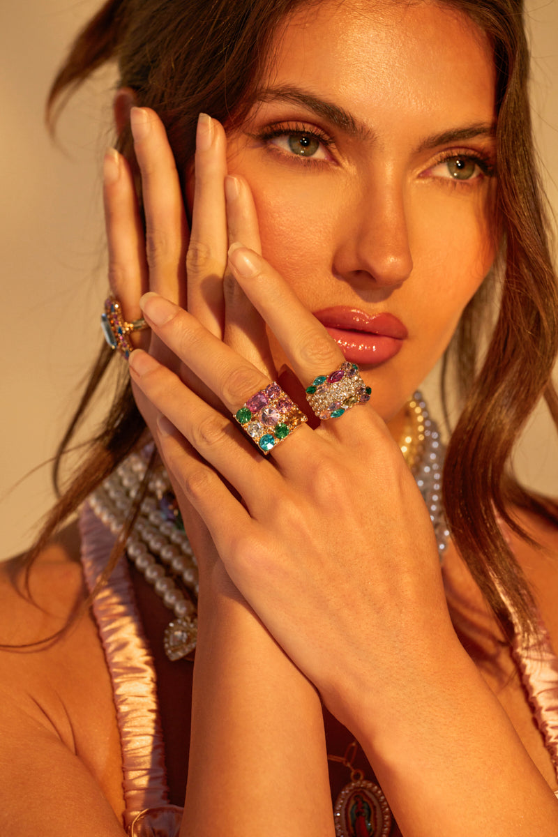 A woman with long brown hair holds her hand to her face, showcasing a stunning THREE WISHES RING SET adorned with colorful jewels. She wears a pearl necklace and a pink top under warm, golden lighting that enhances the brilliance of the sparkling rings on her fingers.
