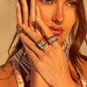 A woman with long brown hair holds her hand to her face, showcasing a stunning THREE WISHES RING SET adorned with colorful jewels. She wears a pearl necklace and a pink top under warm, golden lighting that enhances the brilliance of the sparkling rings on her fingers.