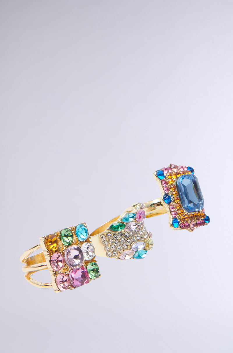 The THREE WISHES RING SET from the AKIRA Label shines on a light gray background. It features three distinct sections adorned with colorful jewels, including blue, pink, green, and clear stones arranged in intricate patterns.