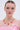 A woman with slicked-back hair models the PICK ME RED NECKLACE, featuring gold rhinestones and dangling red heart charms, paired with a pink off-the-shoulder ruffled top and ribbon bow, posing against a plain light background.