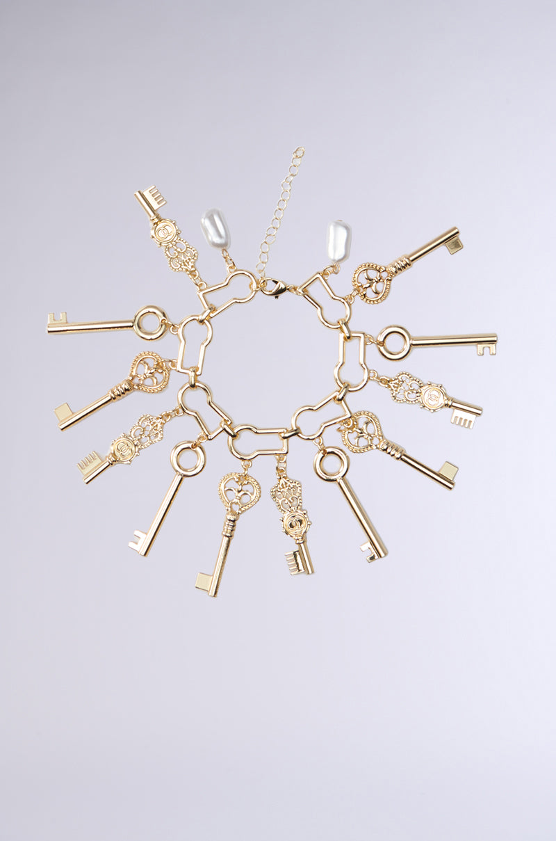 The ALL THE KEYS CHARM BRACELET features classic gold key pendants, lock-shaped hardware, and two pearl-like charms, all elegantly arranged on a neutral light background. 0