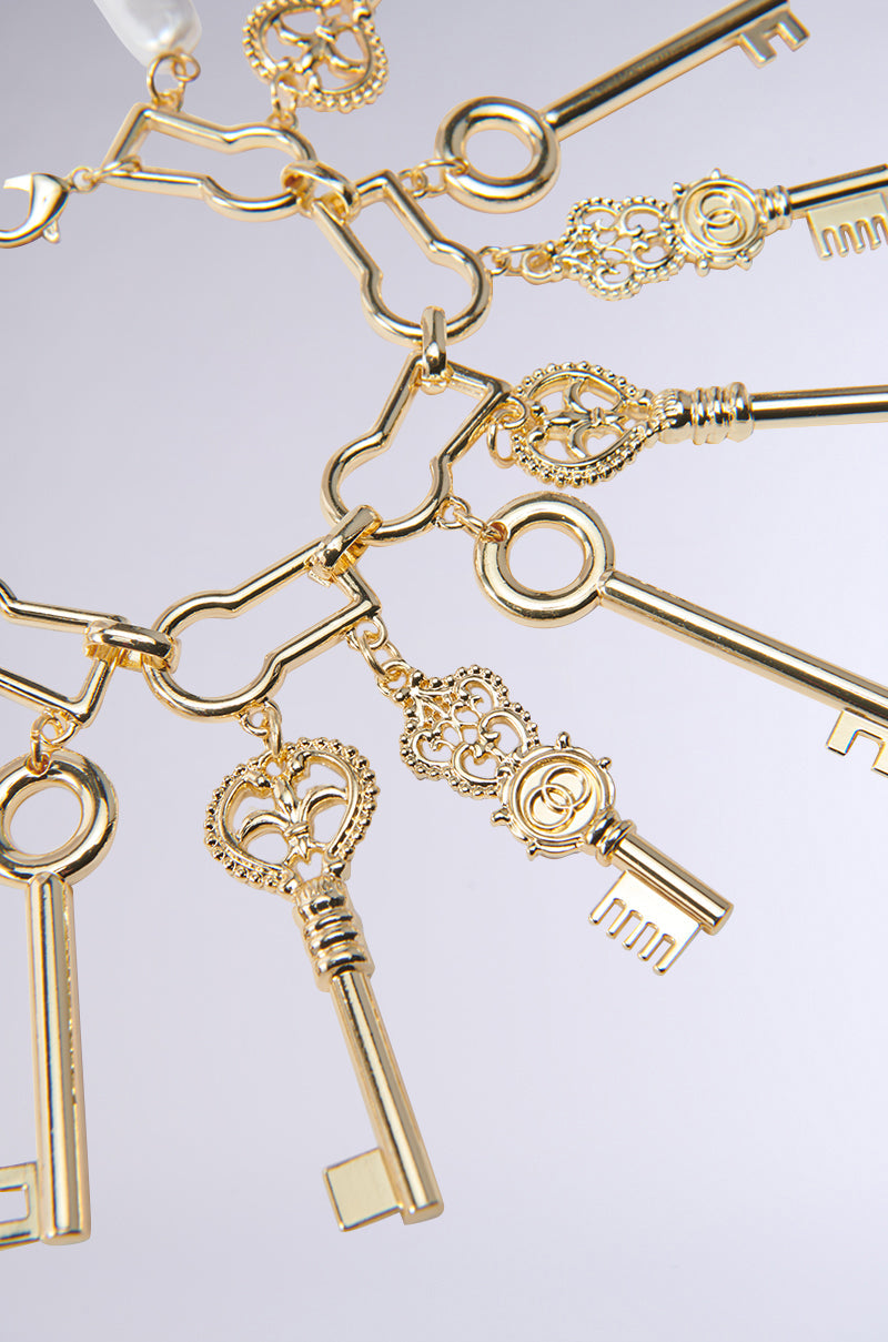 A close-up of the ALL THE KEYS CHARM BRACELET, featuring several ornate, gold-colored vintage key pendants hanging from linked rings and arranged in a circular pattern on a light, reflective background. 3