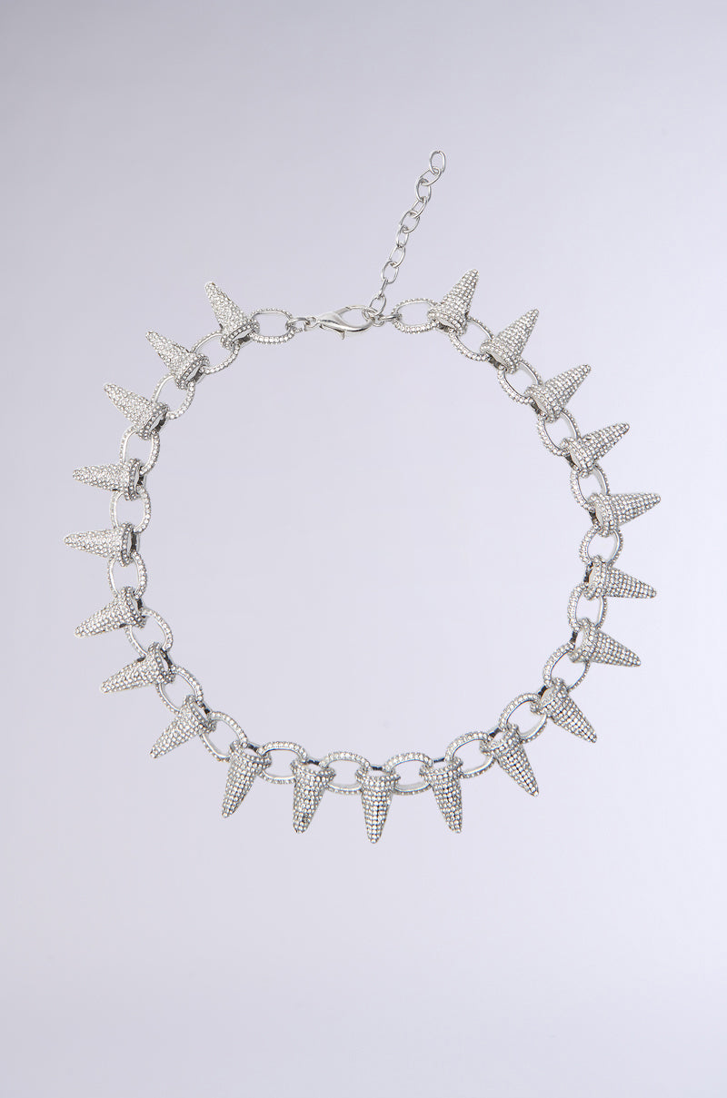 The OH SO LUXE SPIKE NECKLACE is a striking piece featuring silver cone-shaped spikes and rhinestone accents on circular chain links, complete with a clasp and extender chain, beautifully displayed against a light gray background.