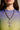 Wearing a blue and green gradient sheer top, the person showcases the BIGGER IS BETTER NECKLACE featuring an oversized cross pendant on a chunky Cuban link chain. Only their lower face, neck, and chest are visible.
