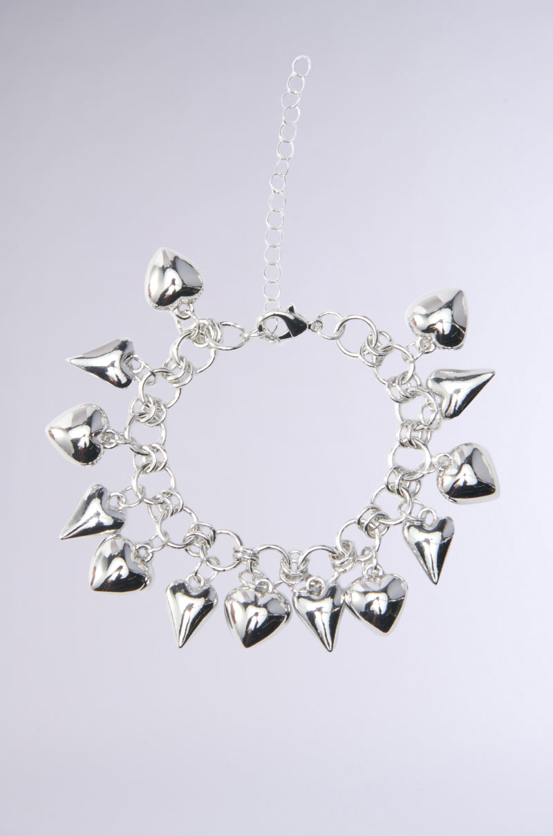 The TOO TRENDY CHARM BRACELET showcases a shiny silver look with several heart-shaped charms and a delicate extension at the clasp. Its chain link design is laid flat on a light gray background. 0