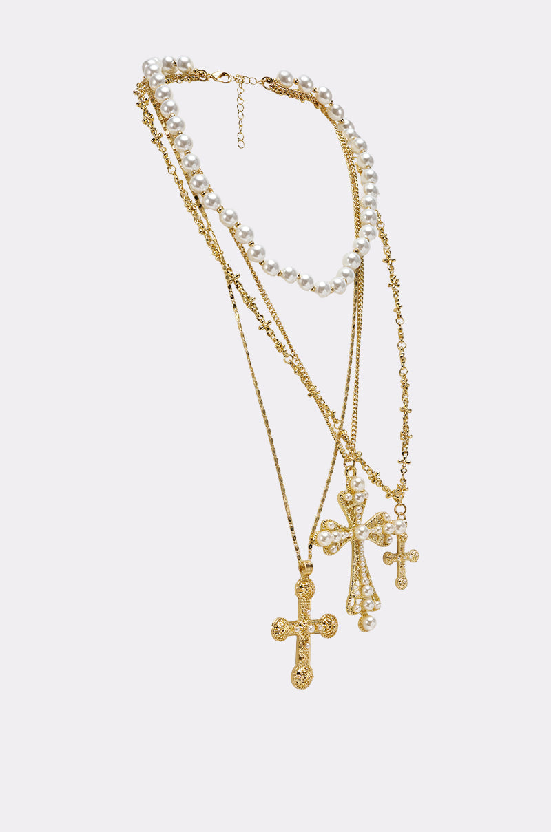 The SIERRA NECKLACE features three layers: a white pearl strand and two gold chains, each with ornate baroque cross pendants, beautifully arranged against a light background. 2