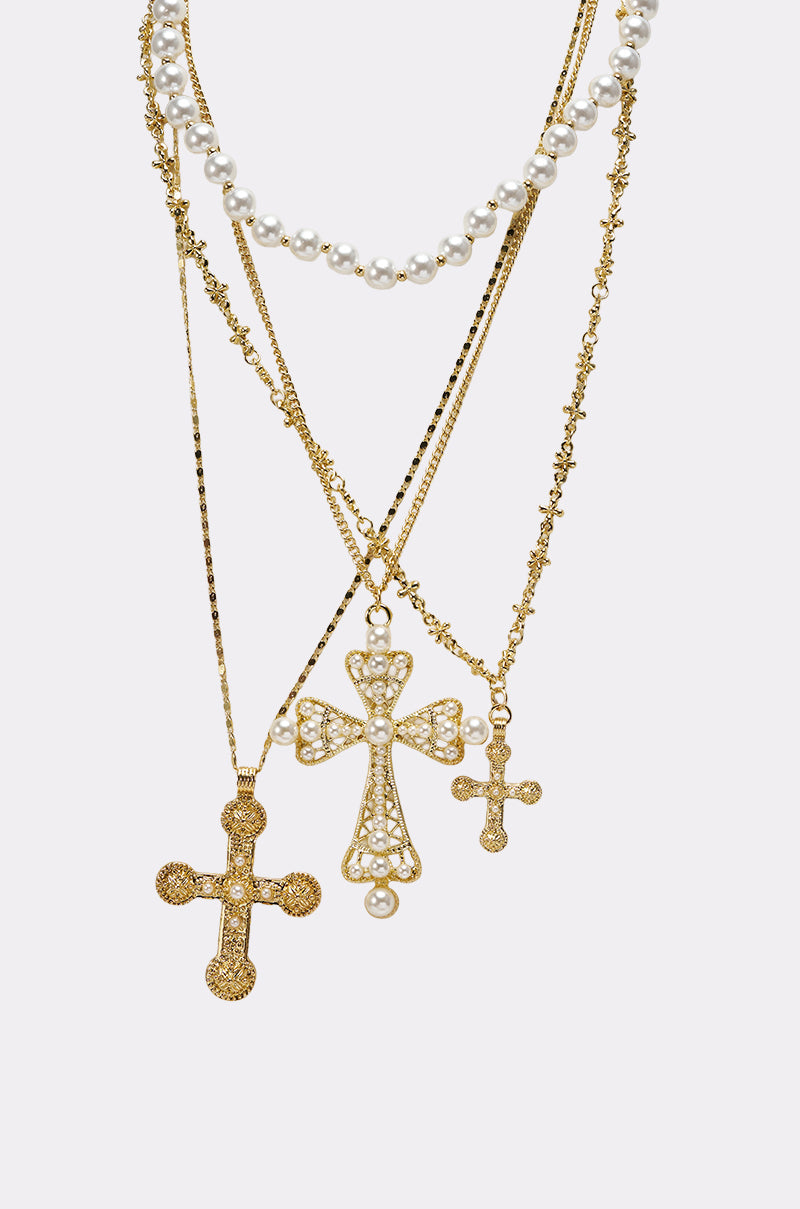 The SIERRA NECKLACE features three layered gold chains with ornate cross pendants and a faux pearl necklace, adorned with intricate designs and small gemstones, styled together against a plain white background. 4