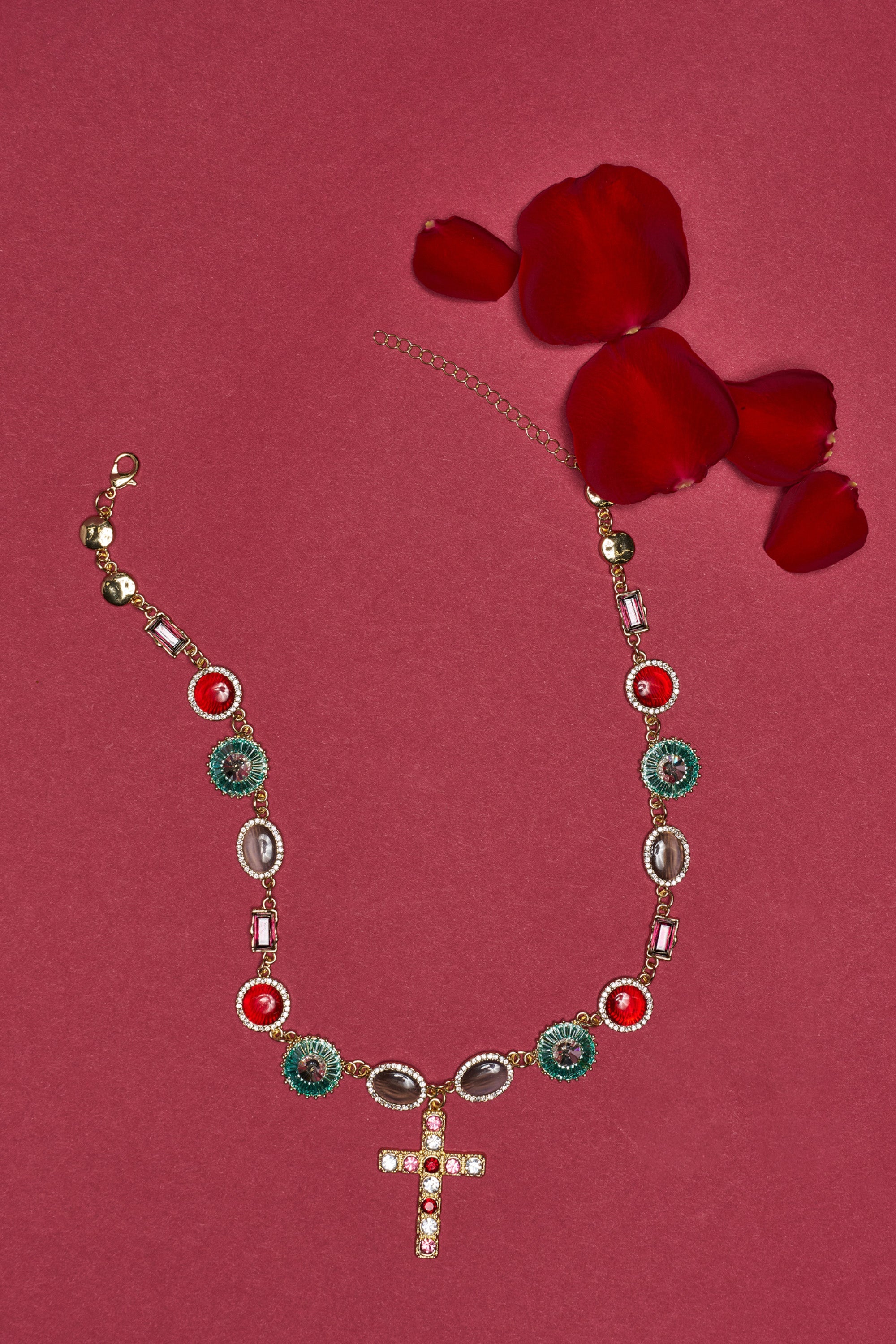 The LUXE TRINITY NECKLACE, featuring a jeweled cross pendant, is displayed on a red surface surrounded by four scattered red rose petals. 1