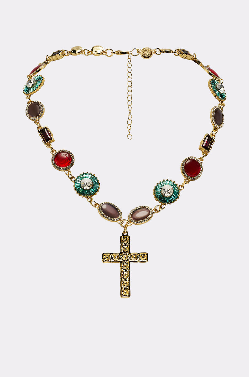 The LUXE TRINITY NECKLACE is a gold-tone piece with colorful oval gemstones and a large cross pendant embellished with small crystals, displayed on a plain white background. 0