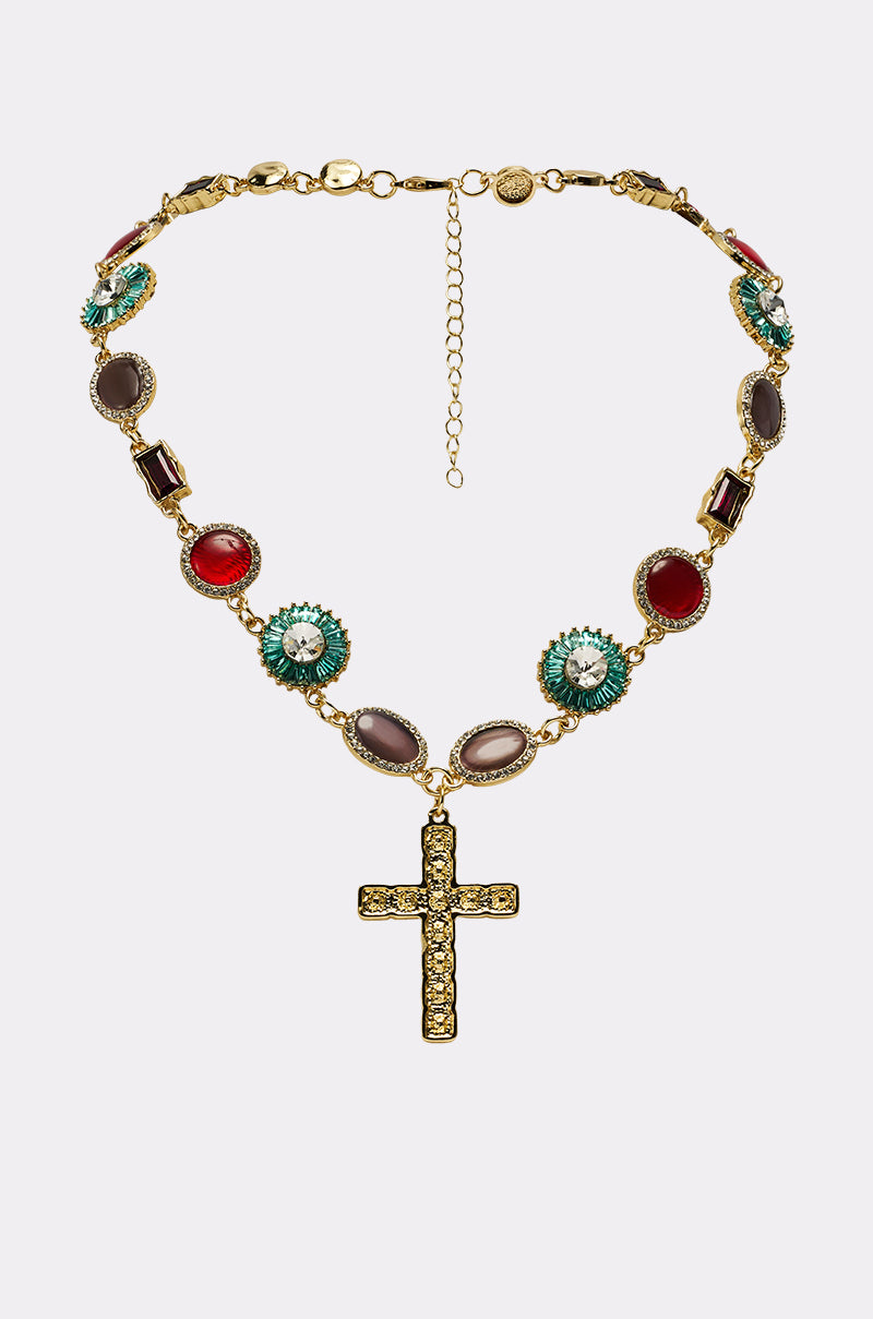 The LUXE TRINITY NECKLACE is a gold-tone piece with colorful oval gemstones and a large cross pendant embellished with small crystals, displayed on a plain white background.