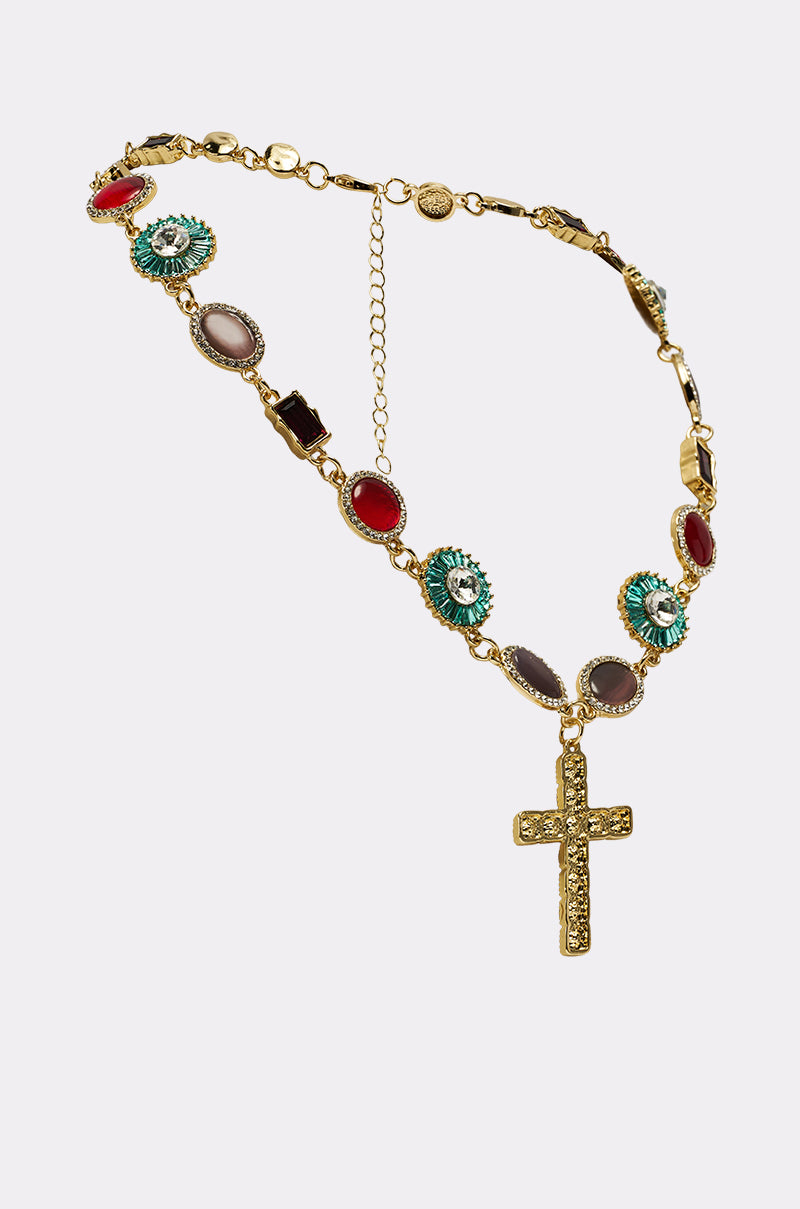 The LUXE TRINITY NECKLACE is a gold-tone piece with colorful oval and round gemstones, highlighted by a striking gold cross pendant embellished with crystals, shown on a plain white background.