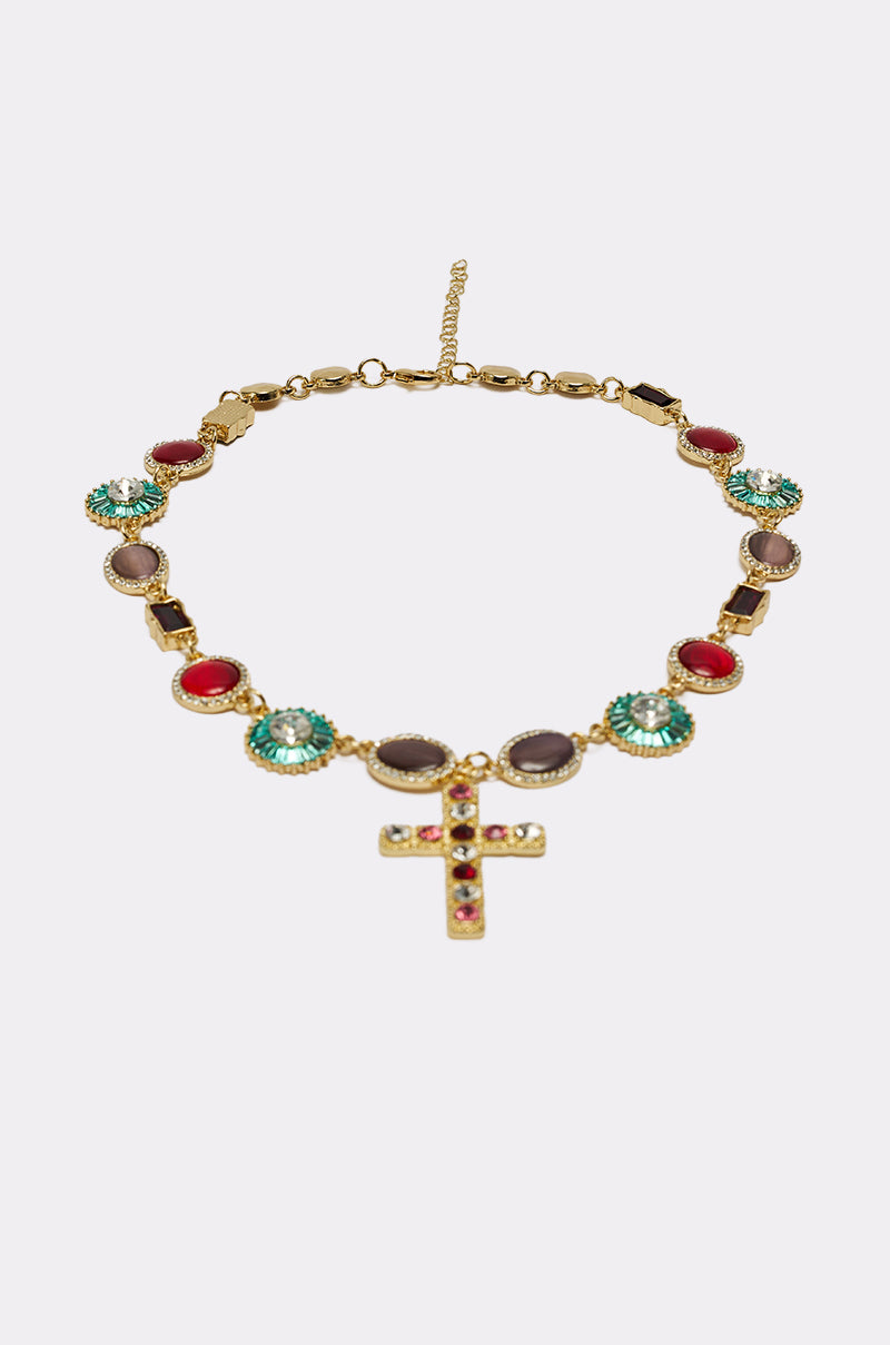 The LUXE TRINITY NECKLACE is a gold-tone piece with alternating round red, turquoise, and brown stones, featuring a cross pendant adorned with red and turquoise gems, shown on a plain white background.