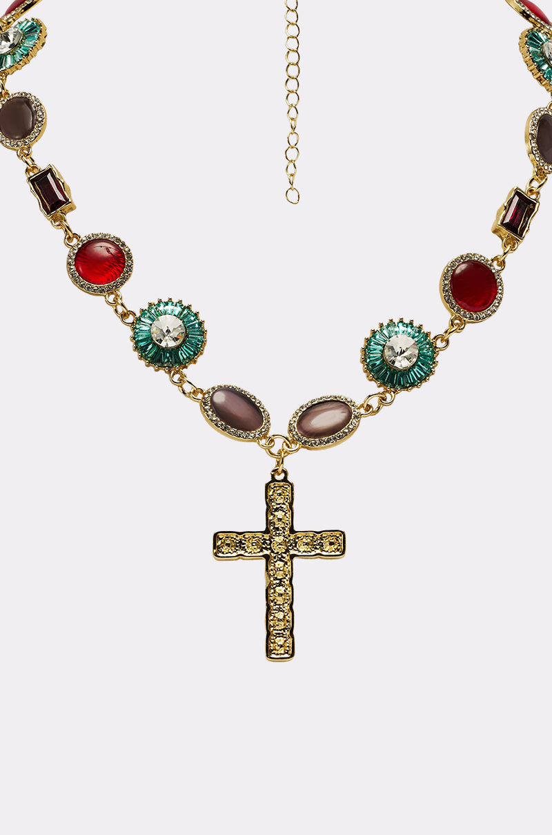 The LUXE TRINITY NECKLACE is a gold-tone piece with a jeweled cross pendant featuring alternating oval and round gemstones in red, green, and neutral shades, each framed by sparkling crystals.