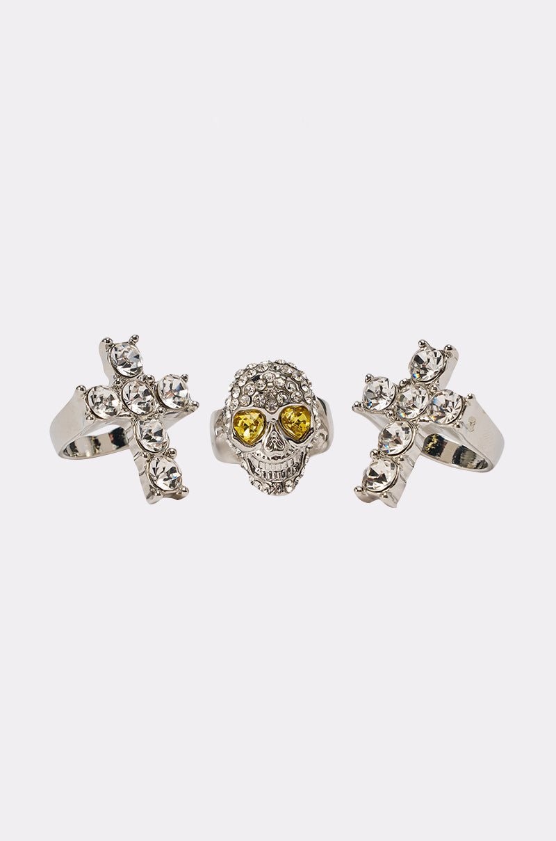 The FOREVER PROTECTED RING SET includes three silver-tone rings: two feature crystal crosses, while the third is a bold skull ring with clear crystals and yellow gemstone eyes, displayed on a plain white background. 1