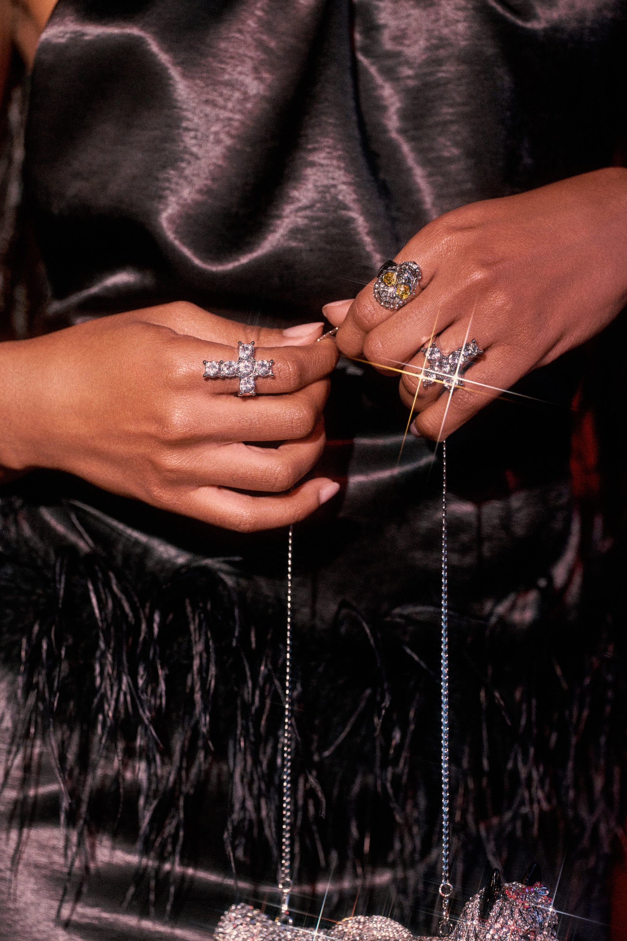 A person in a shiny black feathered dress holds a sparkling handbag by its chain, flaunting the FOREVER PROTECTED RING SET along with bold silver-tone jewelry on both hands. 0