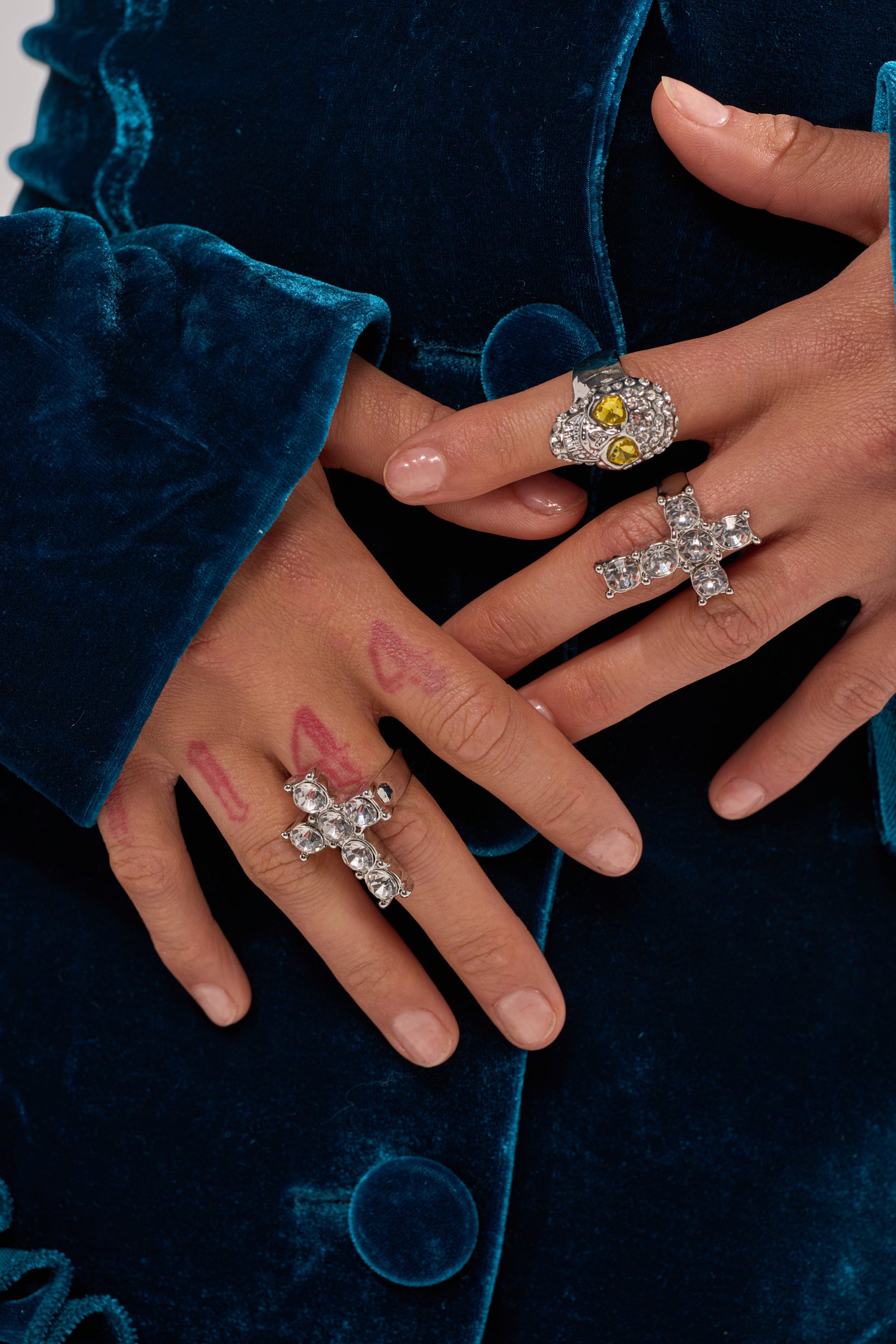 Close-up of hands wearing the FOREVER PROTECTED RING SET—multiple large silver cross and skull rings, some crystal-encrusted, with unique designs. Fingers marked "1 4 4" in red ink. The person wears a blue velvet garment. 2