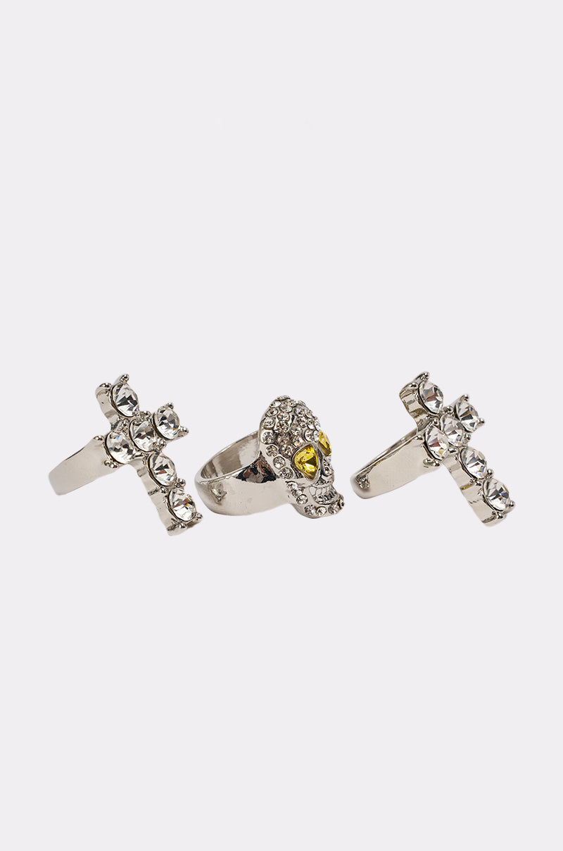 The FOREVER PROTECTED RING SET features three silver-tone rings on a white background: two are crystal cross rings with clear stones, and the center is a skull ring with jeweled yellow gem eyes. 3