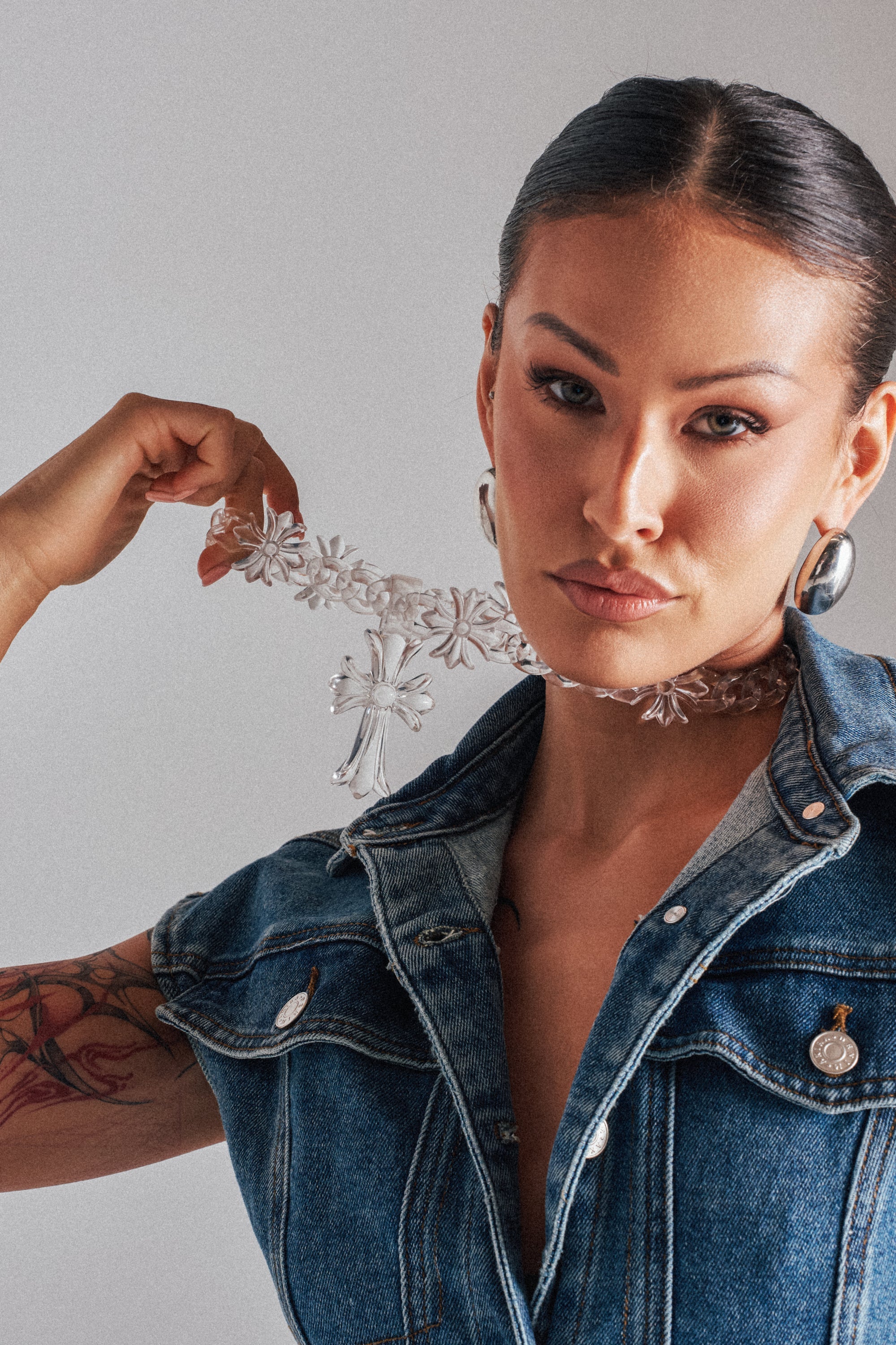 A woman with slicked-back hair, a denim vest, arm tattoo, and silver earrings confidently models the CROSS ME CLEAR NECKLACE—a bold cross pendant on a clear chain—for the camera. 0