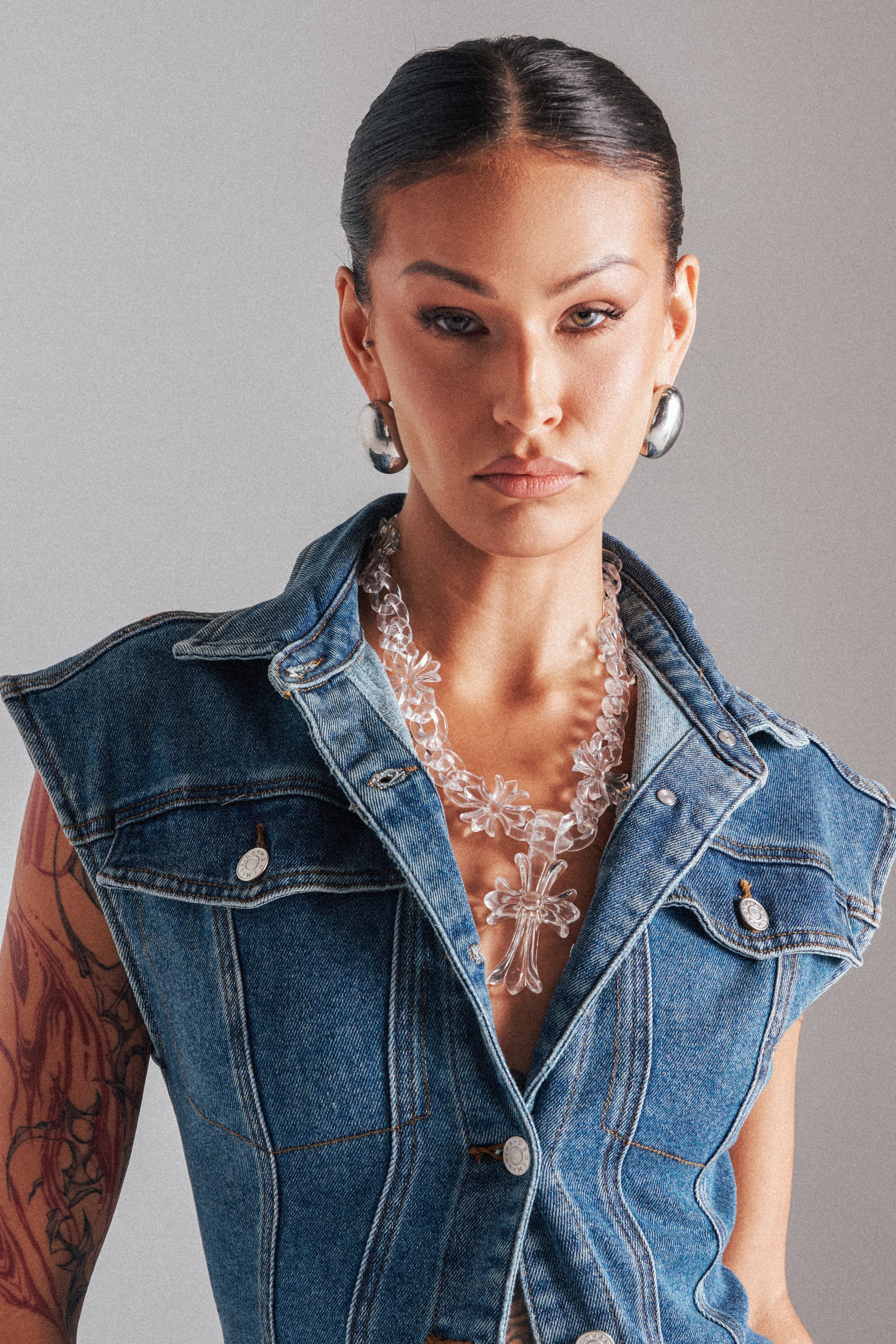 A woman with slicked-back hair wears hoop earrings, statement jewelry including the CROSS ME CLEAR NECKLACE, a sleeveless denim vest, and displays a tattoo on her right arm with a neutral expression. 1
