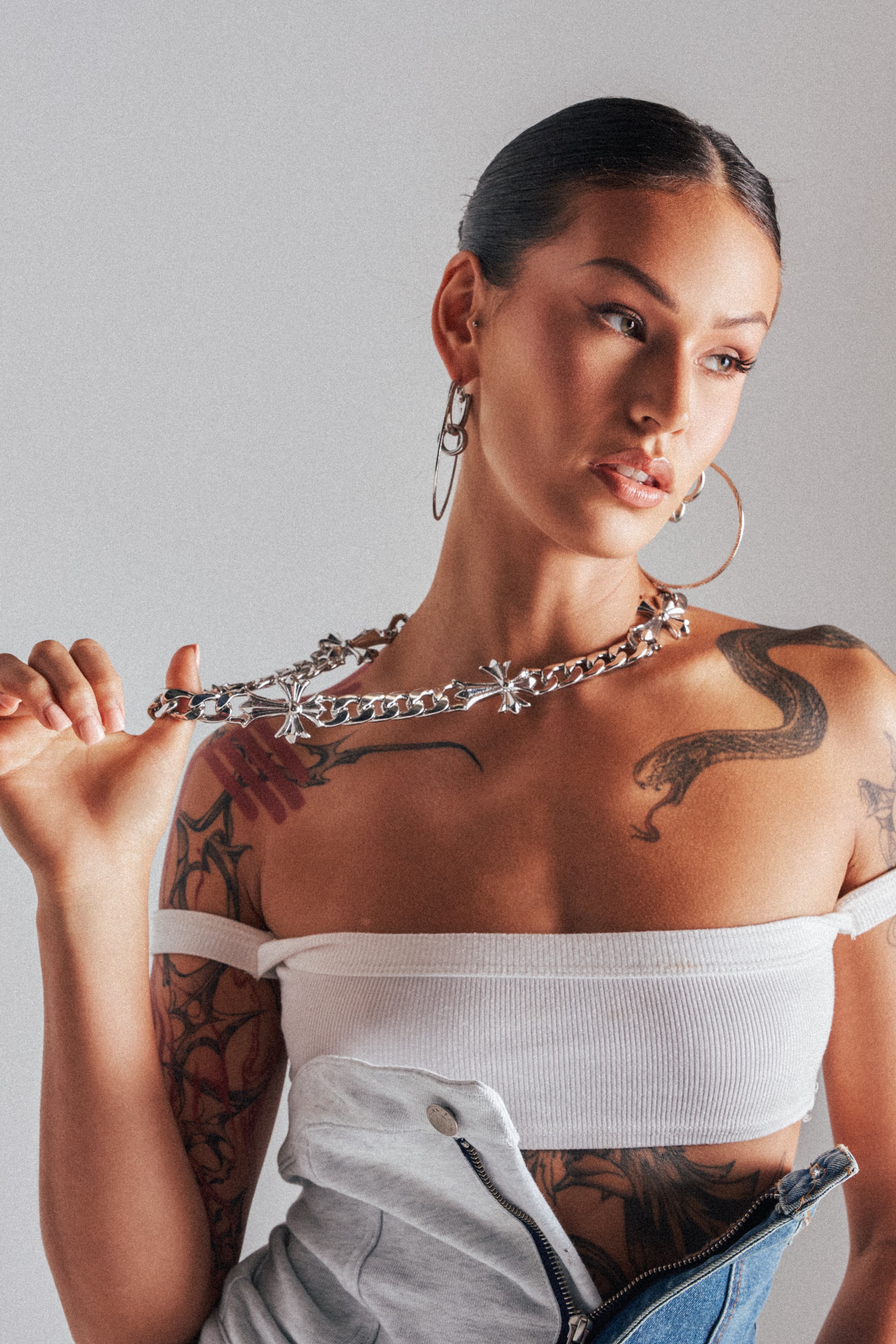 A woman with tattoos and hoop earrings holds the ITS OFFICIAL NECKLACE as a statement accessory. She wears a white off-shoulder crop top and blue jeans, her slicked-back hair showing off a dragon tattoo on her arm.