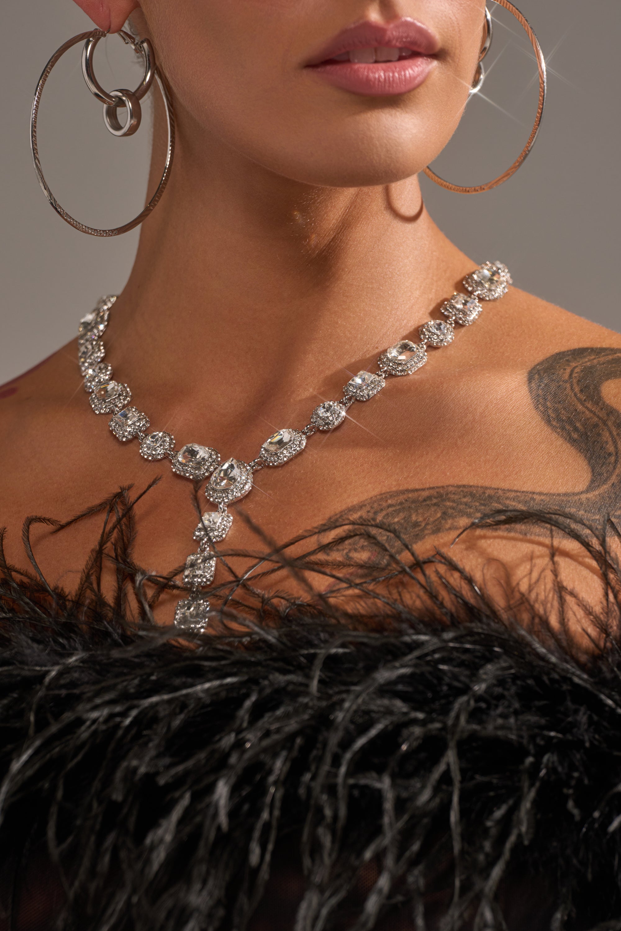 A woman showcases the LUXY LARIAT NECKLACE, paired with large hoop earrings and a black feathered top. A black tattoo accents her shoulder and collarbone, while the image highlights her neck, lips, and striking jewelry.