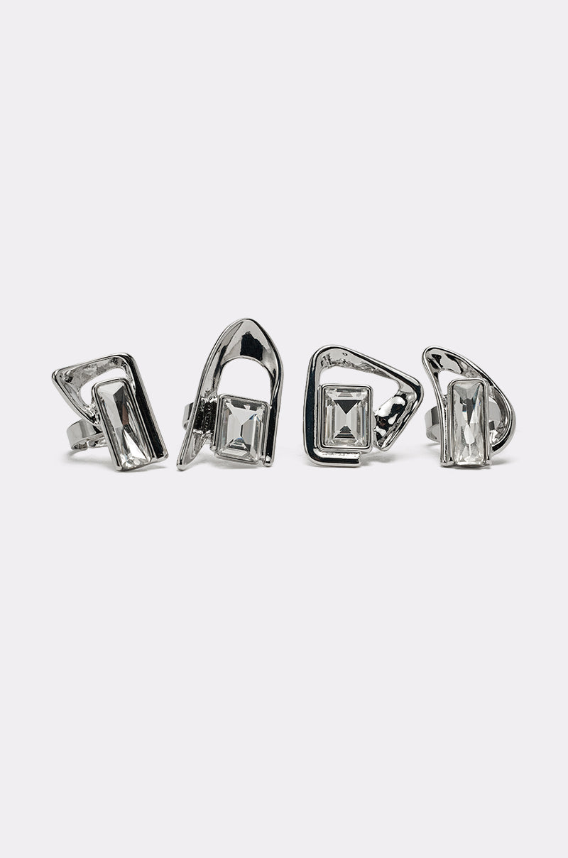 Four geometric rings from the TRUDY SILVER RING SET, each featuring asymmetrical designs and clear rectangular stones, are displayed upright in a row on a plain light background, highlighting this striking futuristic jewelry set. 0