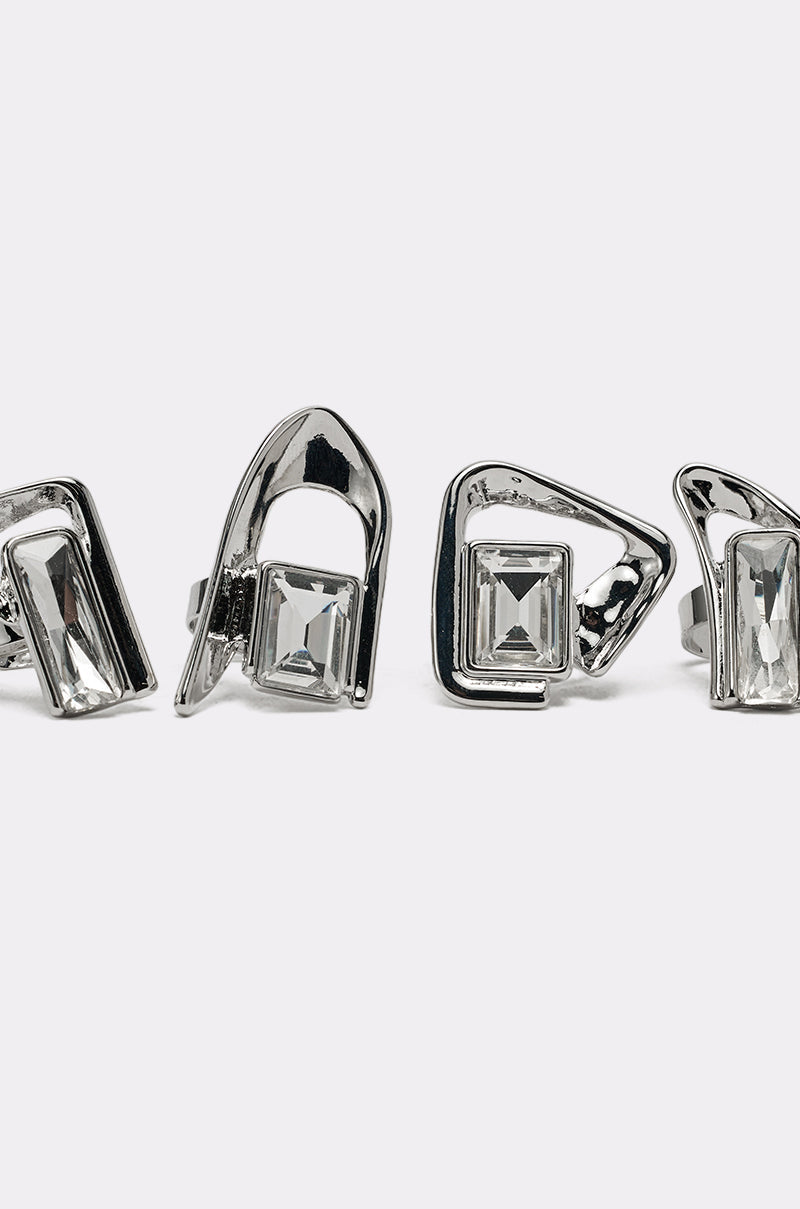 The TRUDY SILVER RING SET features four silver-tone rings with geometric, asymmetrical settings and clear rectangular gemstones, displayed against a plain white background for a modern look. 6