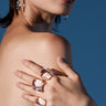 A woman wearing statement earrings and the DIRTY MARTINI RING SET poses with her hands on her bare shoulder, dressed in a burgundy lace-up top against a blue background.