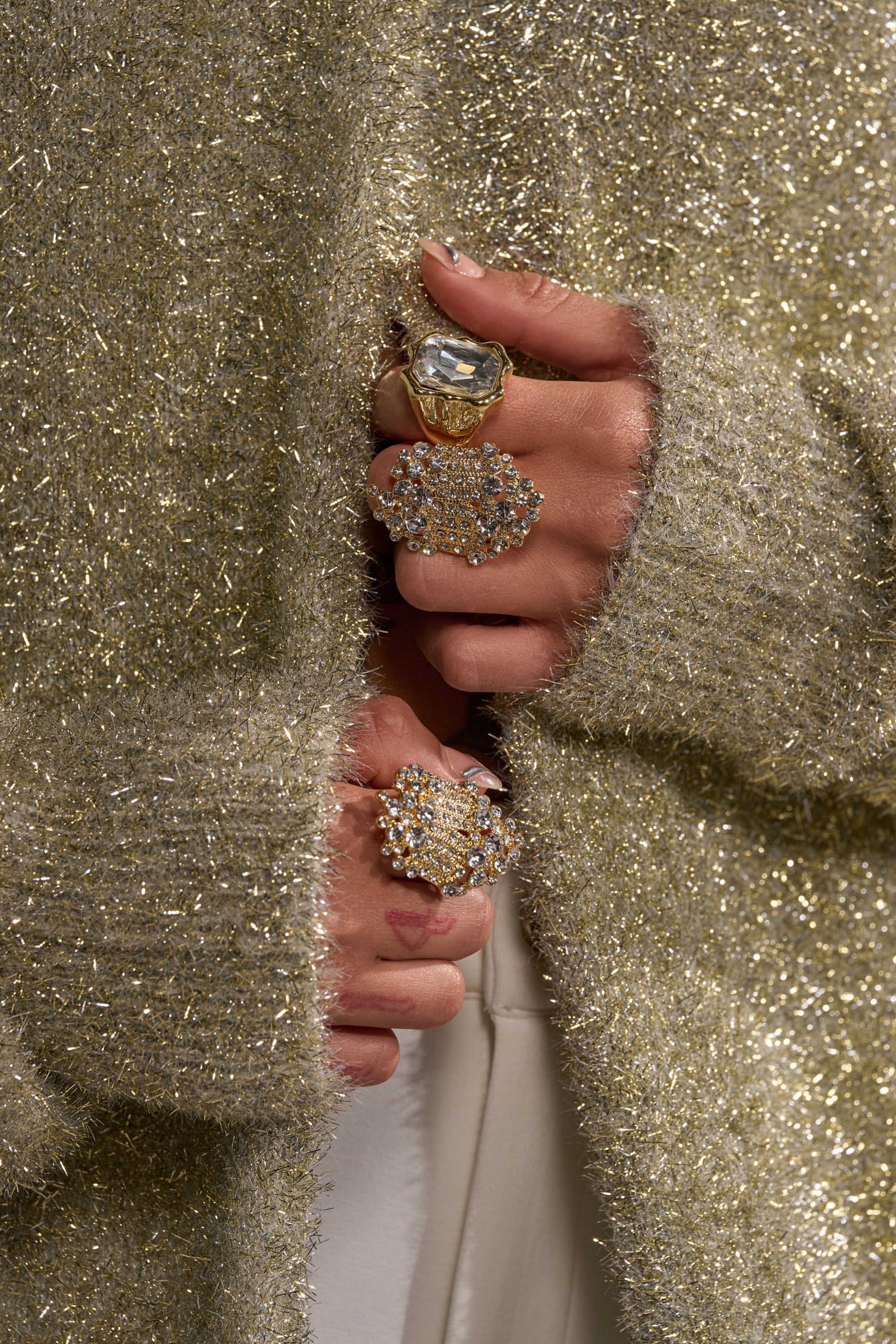 Close-up of hands wearing the CAN YOU NOT RING SET: gold-tone, crystal-embellished rings, partially covered by a glittery gold sweater. Large, ornate rings are paired with natural nails and cream-colored pants. 1