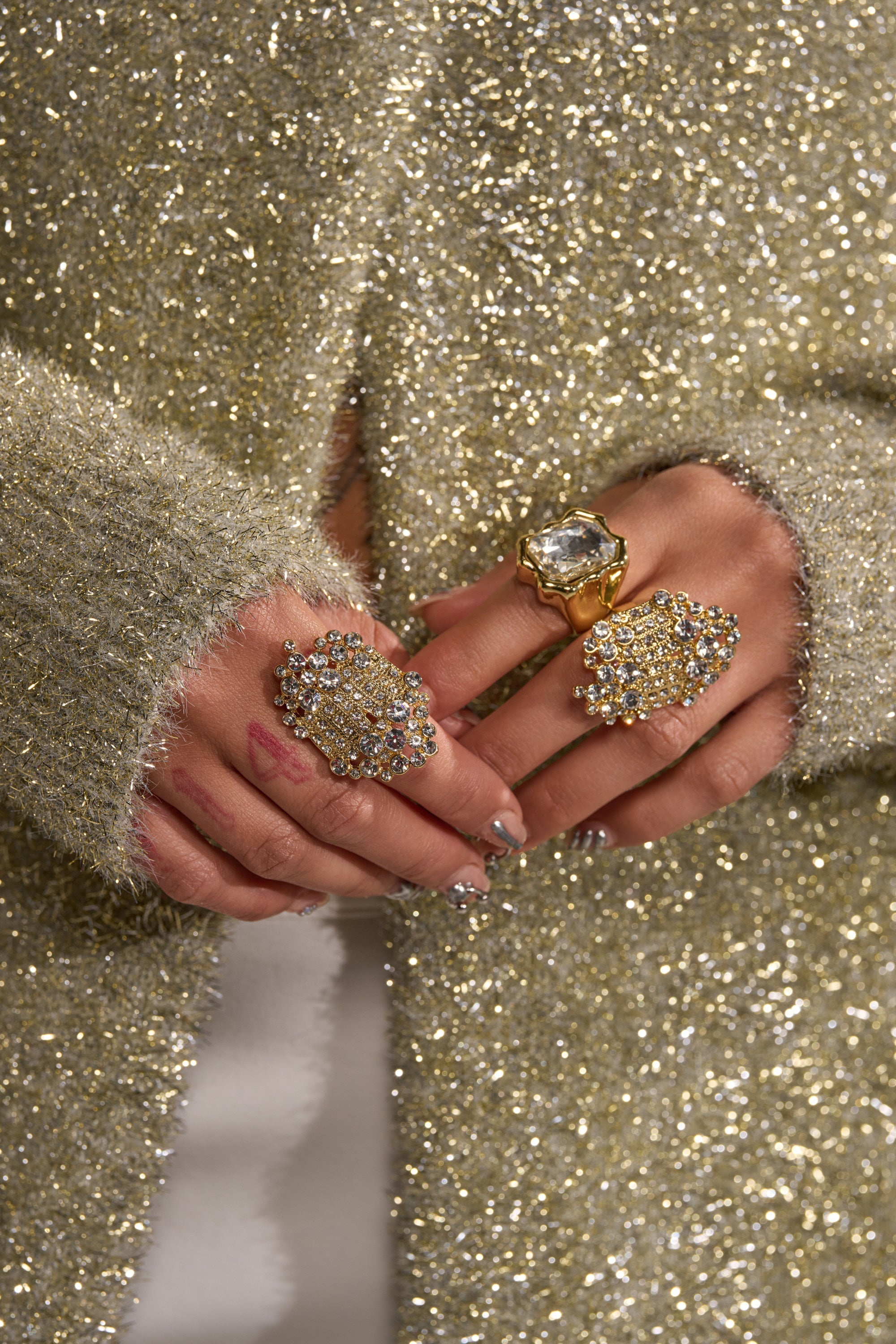 Close-up of hands wearing the CAN YOU NOT RING SET, featuring gold-tone rings with crystal embellishments, paired with metallic nails and subtle finger tattoos against a glittery gold sweater for a standout look. 5