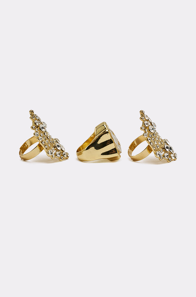 The CAN YOU NOT RING SET features three adjustable gold rings with elongated, textured designs and embedded clear gemstones, shown from the front, side, and back on a plain white background. 2