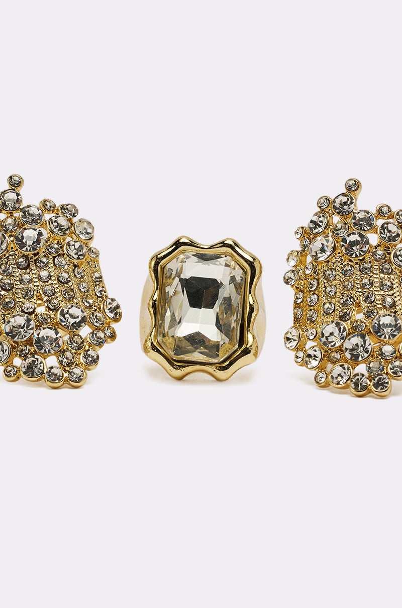 The CAN YOU NOT RING SET includes three crystal-embellished rings: a center gold ring with a large rectangular gemstone in a wavy setting, flanked by two rings featuring clusters of small round crystals for extra sparkle. 4