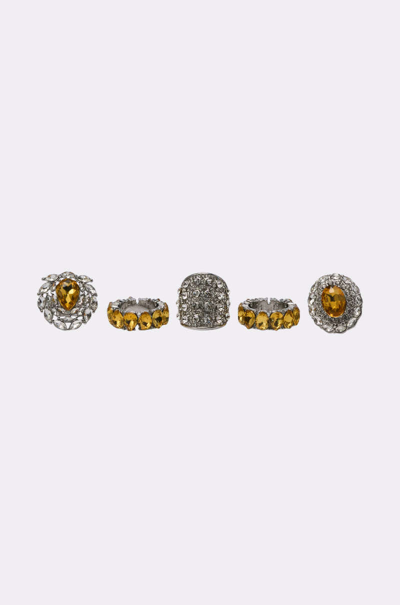 Five ornate rings from the LUXE TIS THE SEASON RING SET are arranged in a row on a white background, showcasing intricate silver designs, yellow gemstones, and a pavé dome ring with sparkling stones at the center. 0