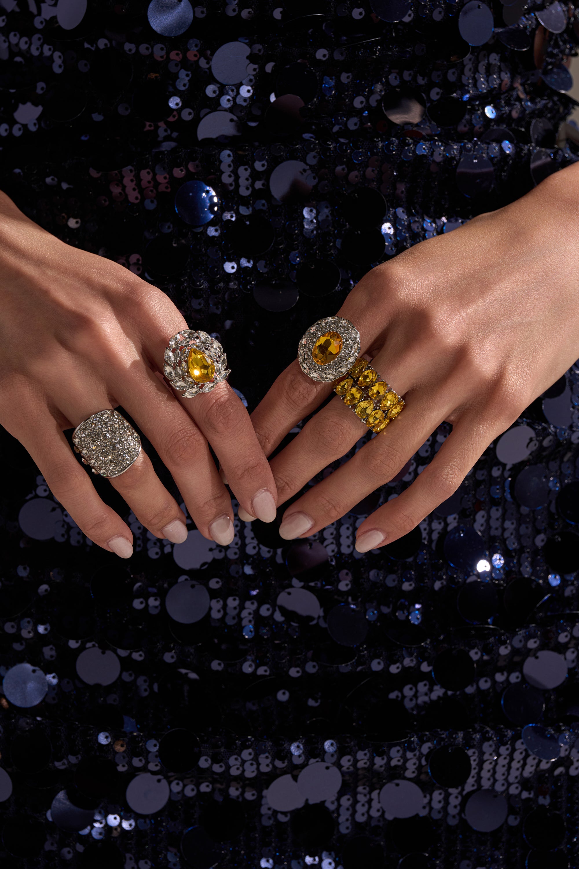 A close-up of two hands wearing the LUXE TIS THE SEASON RING SET with yellow and clear gems; paired with manicured nails and a dark blue sequined dress, it's perfect for a holiday glam rings style.
