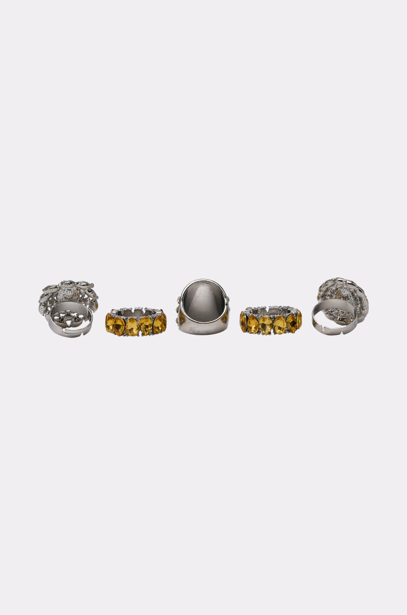 The LUXE TIS THE SEASON RING SET showcases five silver rings on a white background, featuring ornate designs: two with yellow gemstones and three detailed cocktail rings with intricate silver patterns for holiday glam. 2