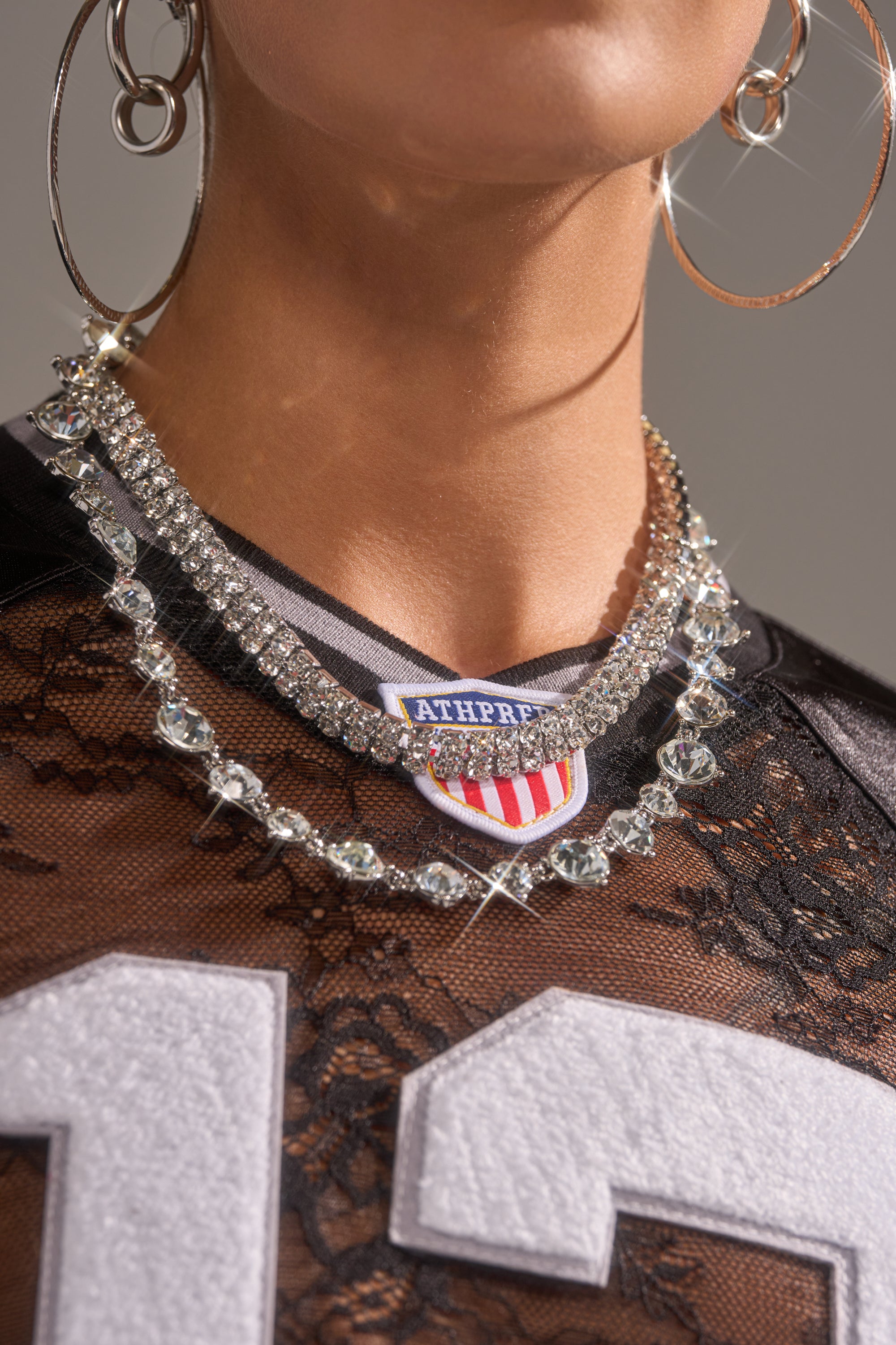 A person wearing large hoop earrings, the LOVER NECKLACE, a rhinestone layering necklace, a black lace top, and a sporty jersey with the number 13 and an "ATHPREP" badge on the collar.