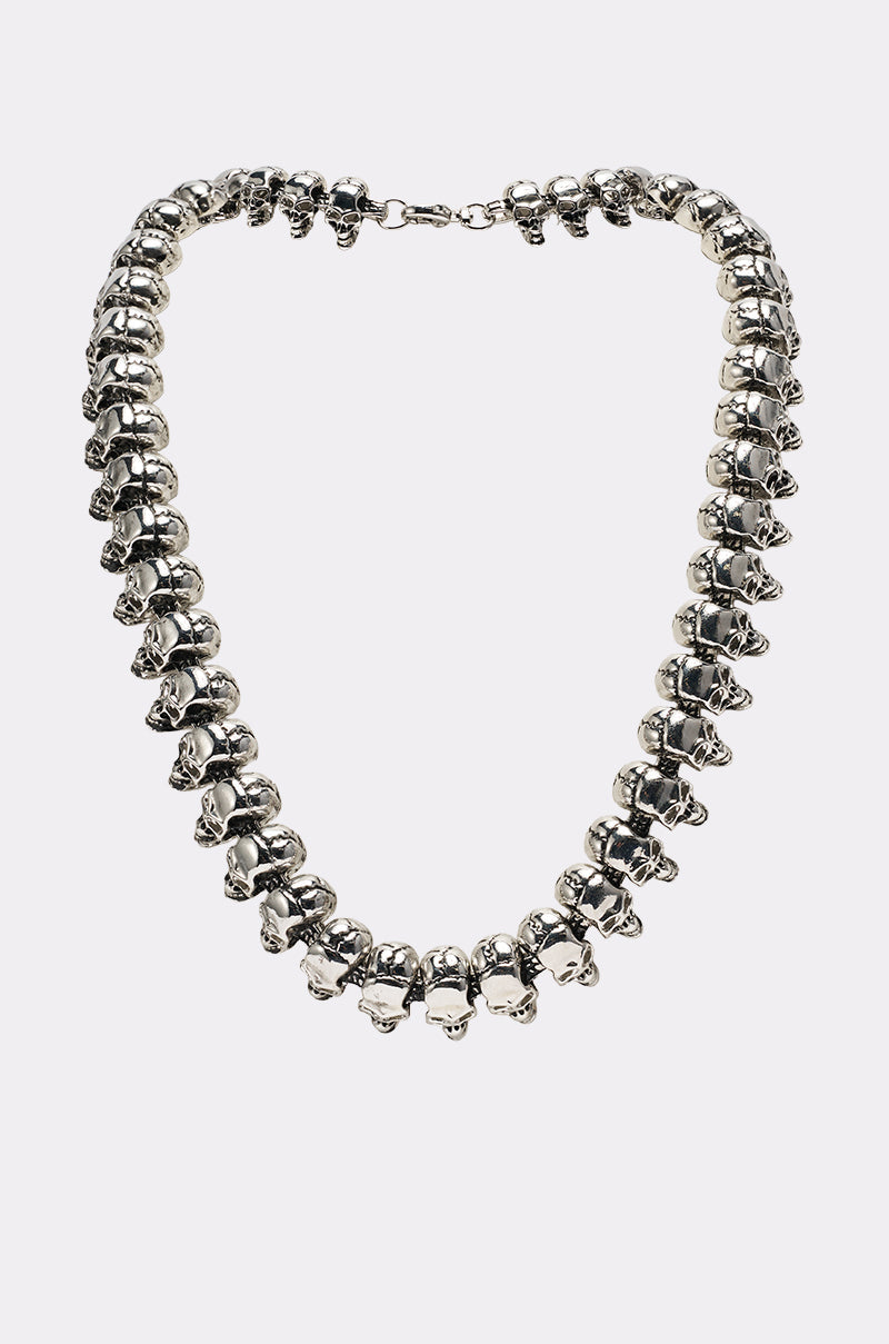 The SKULLY NECKLACE is a silver chain featuring small, evenly spaced skull-shaped beads linked together to form a complete loop, secured with a clasp and shown against a plain light background. 1