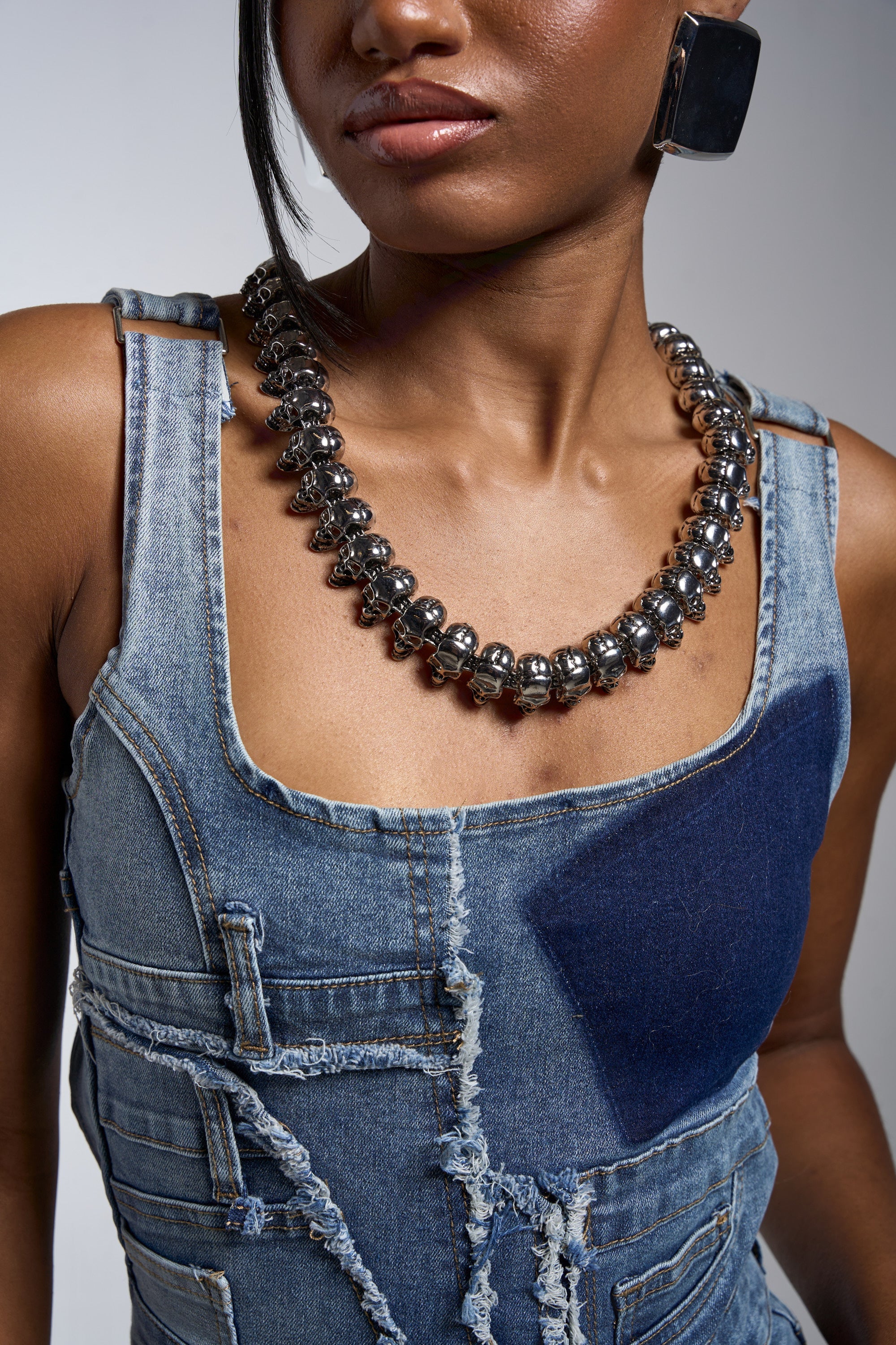 A woman wears a frayed denim overall dress, large square earrings, and the SKULLY NECKLACE. Her face is partially visible, and her hair is styled to one side. 2