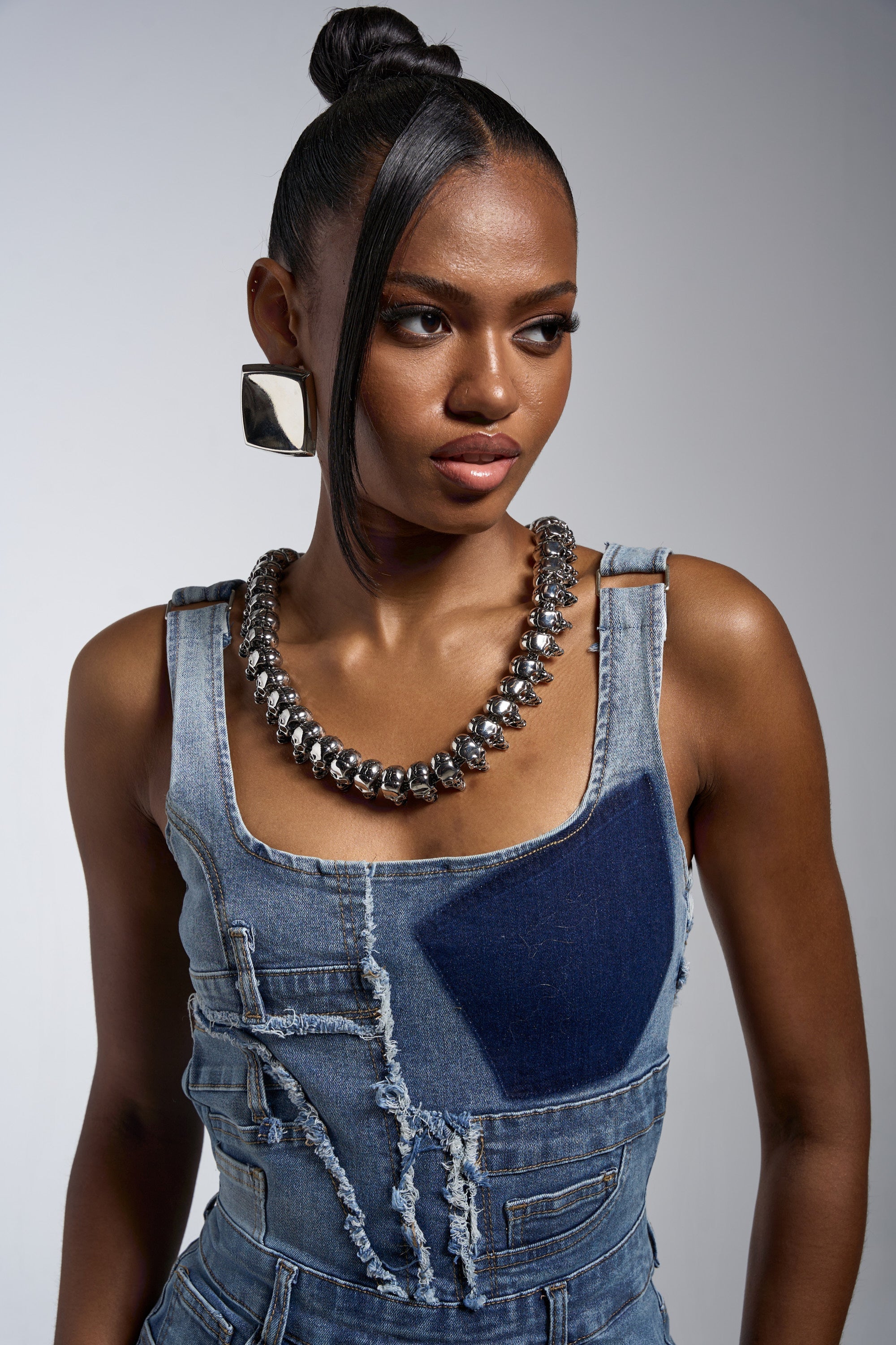 A woman in a patchwork denim top and large square earrings looks to the side, wearing the SKULLY NECKLACE. Her hair is in a sleek bun with a face-framing strand, set against a plain, neutral background. 3