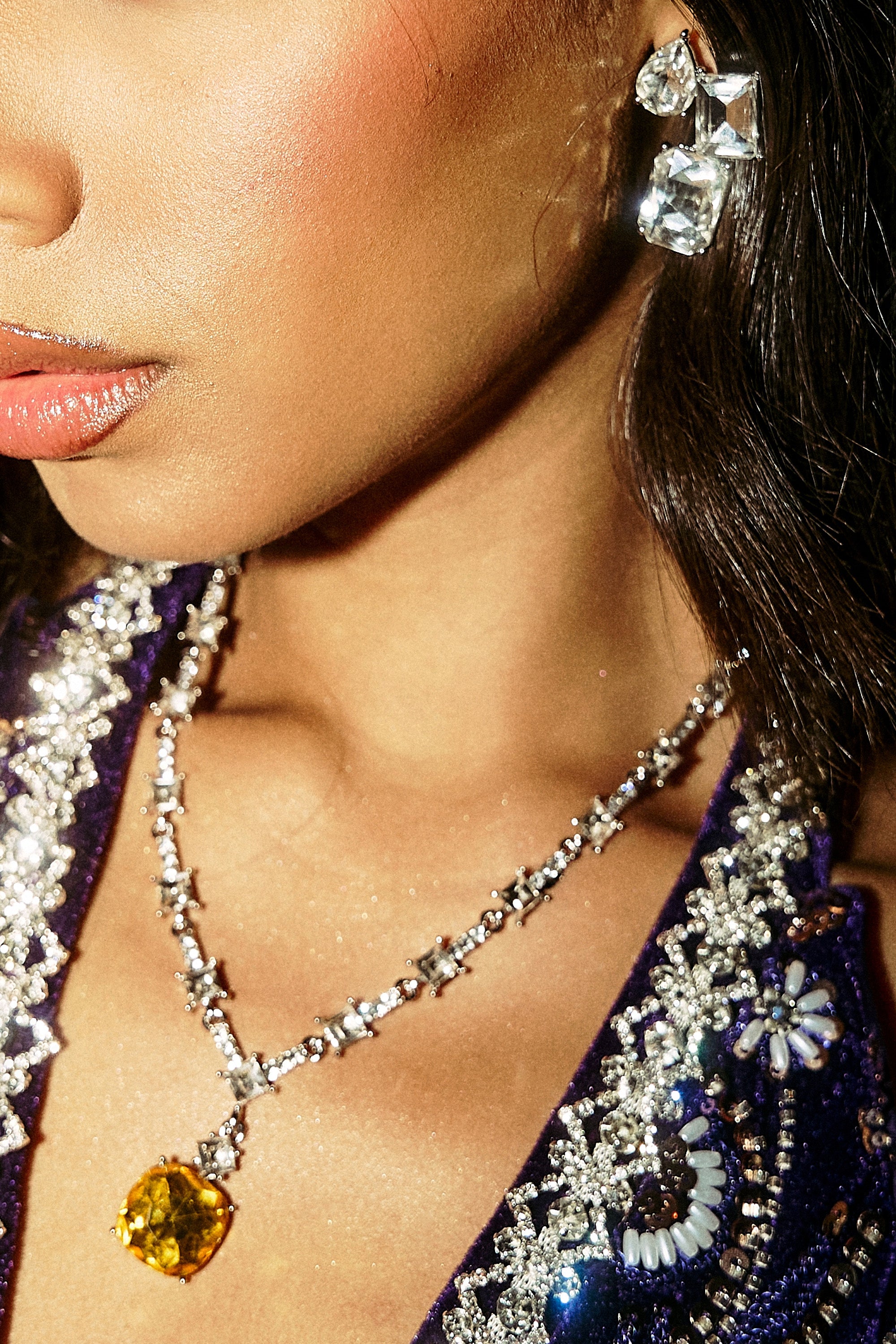 A woman wears sparkling crystal earrings and the LUXE KATE NECKLACE, featuring a bold yellow gemstone, paired with a sequined purple beaded outfit. Only part of her face and upper chest are visible in this close-up.