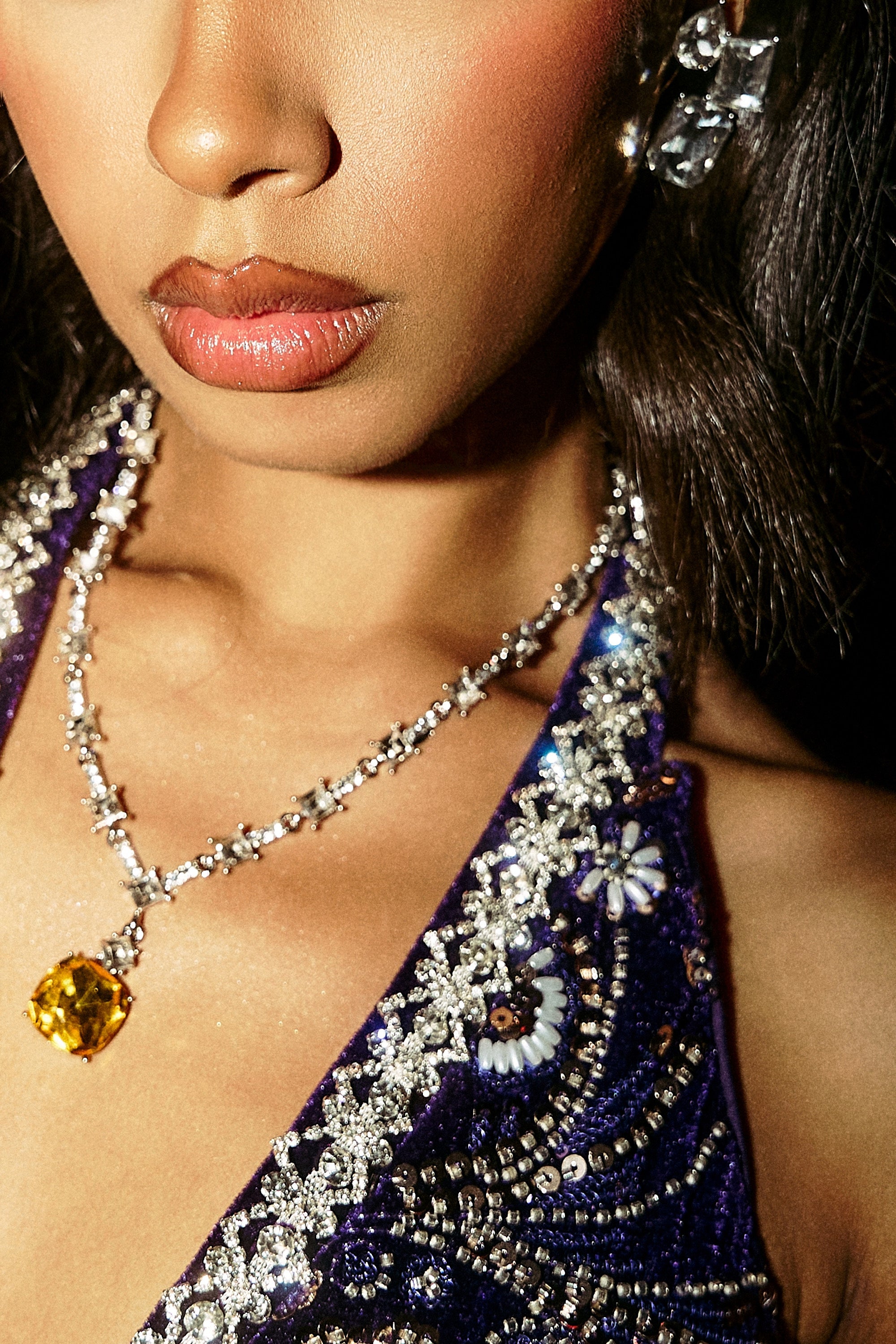 A close-up of a woman in a sparkling purple dress with intricate beading, crystal earrings, and the LUXE KATE NECKLACE—a silver crystal necklace featuring a large yellow gemstone pendant. Only her lower face and upper chest are visible.