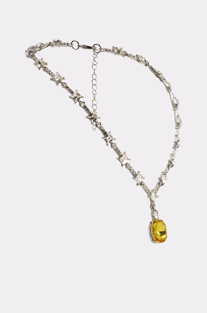 The LUXE KATE NECKLACE showcases an asymmetrical silver design with star accents, square-cut crystals, and a vibrant yellow gemstone pendant against a white background. 2
