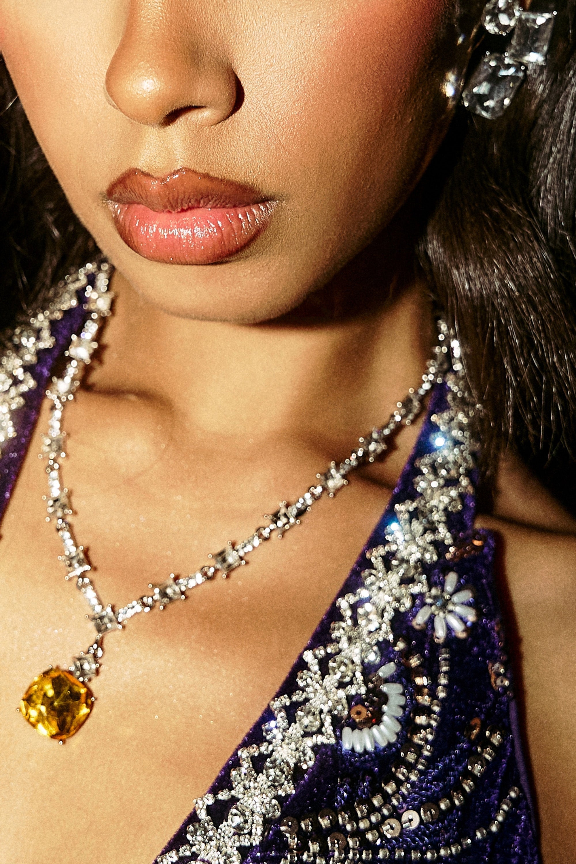 A close-up of a woman wearing the LUXE KATE NECKLACE, featuring a sparkling yellow gemstone pendant, jeweled earrings, and an ornate beaded top. Her lips, nose, and chin are partially visible. 1