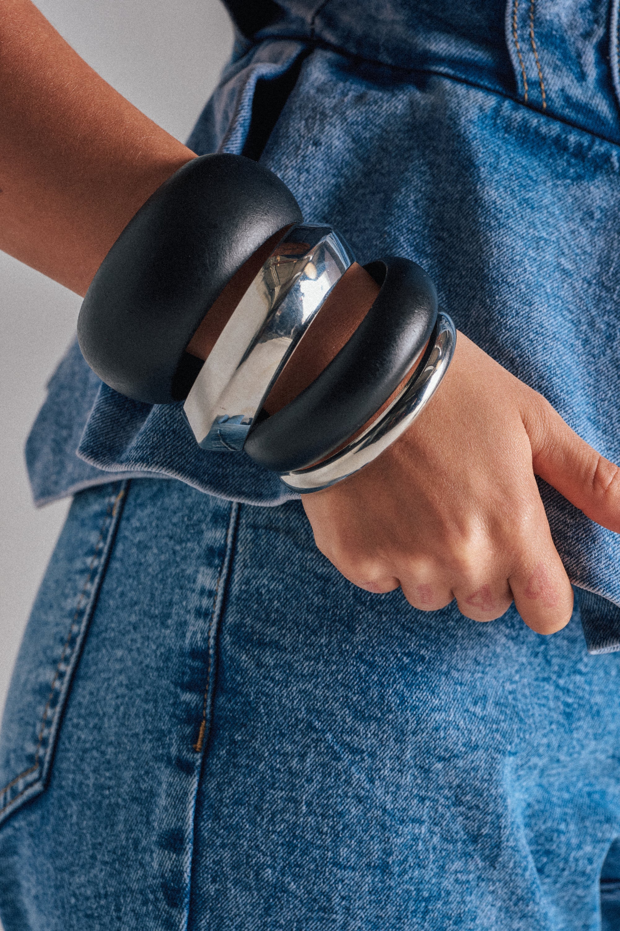 Wearing blue denim jeans and a matching vest, a person stands side-on with their hand on their hip, showcasing the MOD BRACELET SET—featuring chunky black bangles and silver-tone bracelets on their wrist. 0