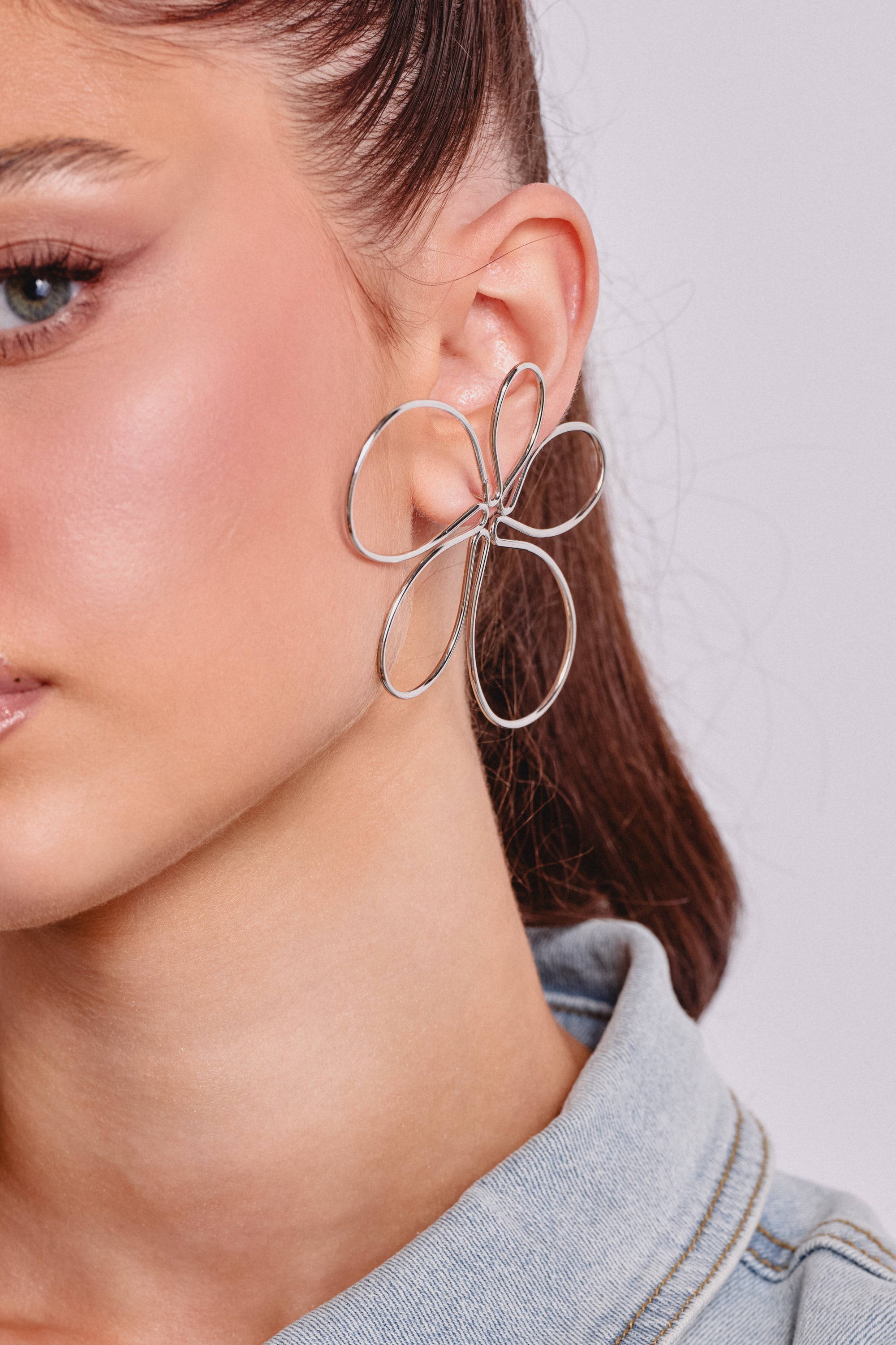 A close-up of a woman's ear showcases the LOVEY EARRING, featuring statement oversized silver-tone wire petals shaped like flowers. 0
