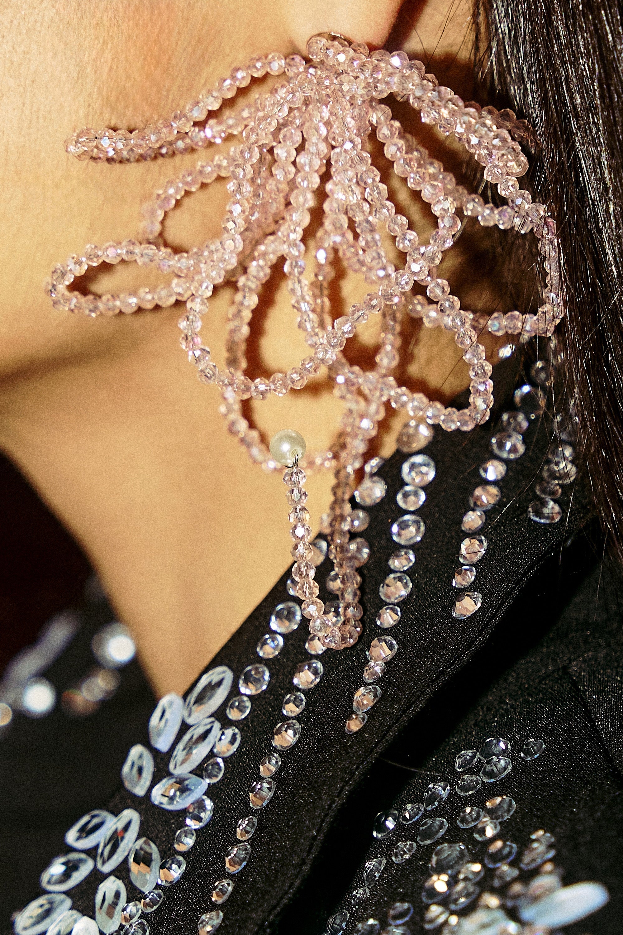 Close-up of a striking GIRL PWR EARRING, a large pink crystal piece shaped like a flower or sea creature, worn on the ear. Paired with a black garment adorned in shiny silver sequins for an eye-catching AKIRA Label look. 3