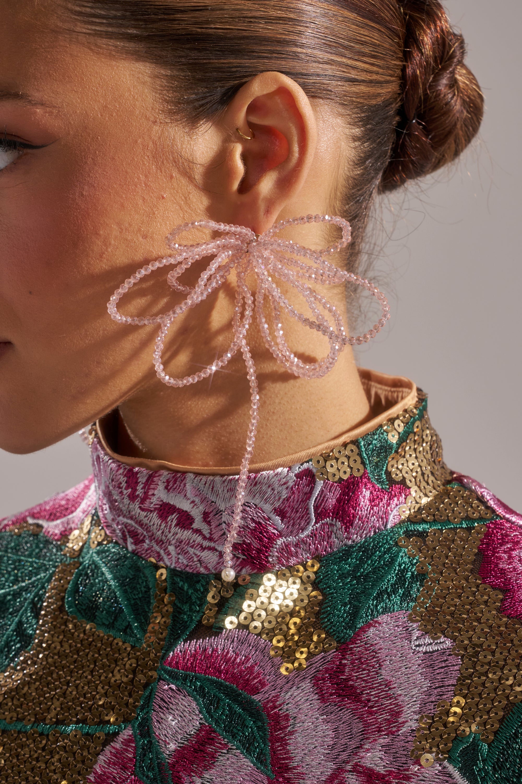 A close-up of a person wearing the GIRL PWR EARRING, styled as large pink floral statement earrings, paired with a gold ear cuff and a high-collared floral sequin top in pink and green; hair is in a neat bun. 1